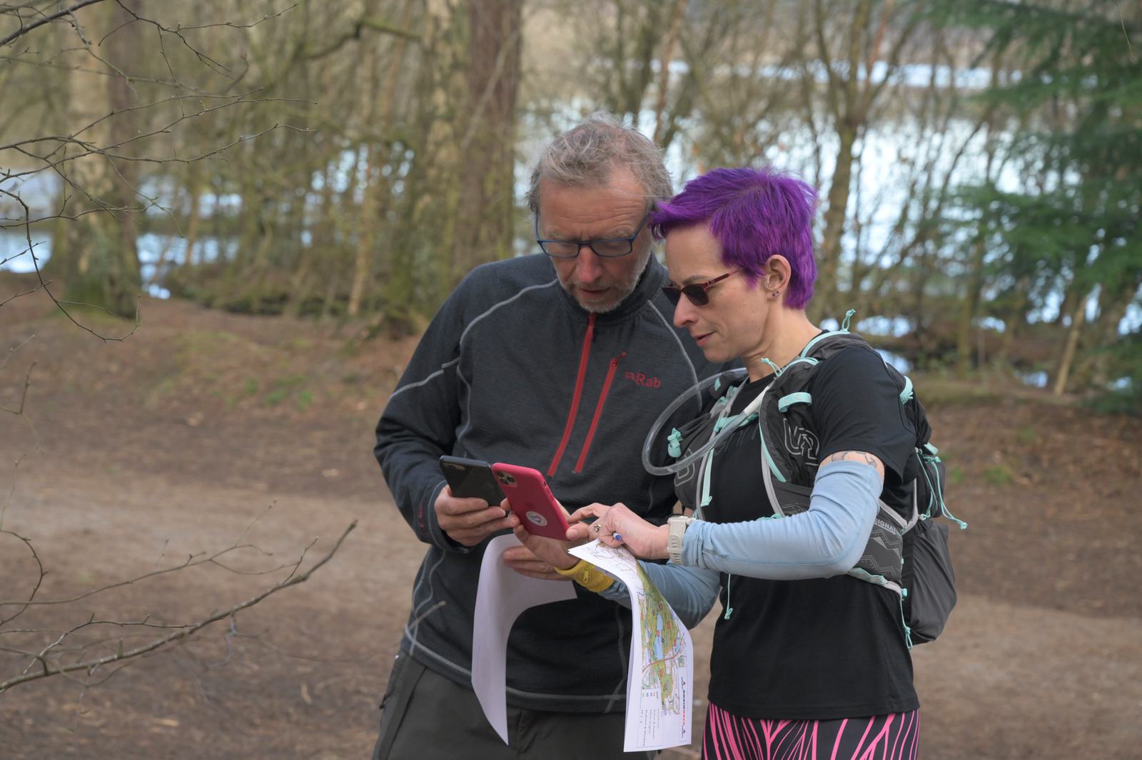 Two people standing outside in a wooded area beside a body of water. They are looking at a map and a smartphone. The person on the right is wearing a hydration backpack and has short purple hair. Both appear to be focused on navigating the trail.