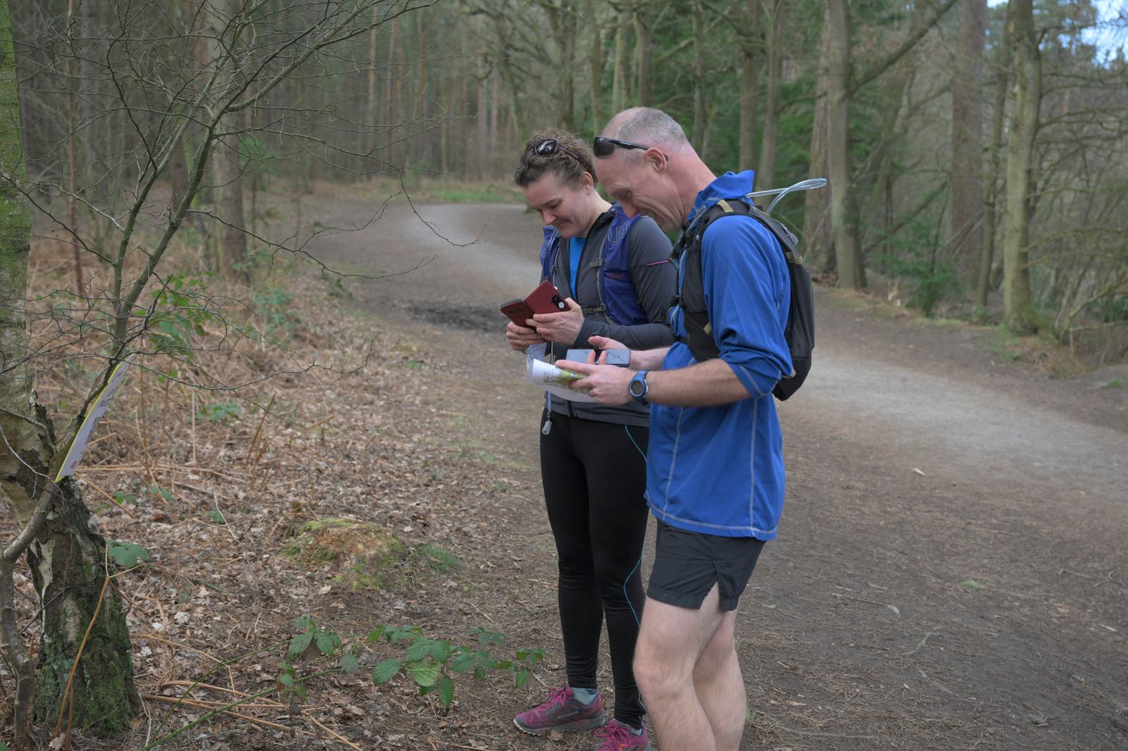 Two people dressed in athletic wear stand on a forest trail. They both appear to be checking maps or devices. Trees and foliage surround them, and a dirt path extends into the forest behind them. One person wears a hydration pack.