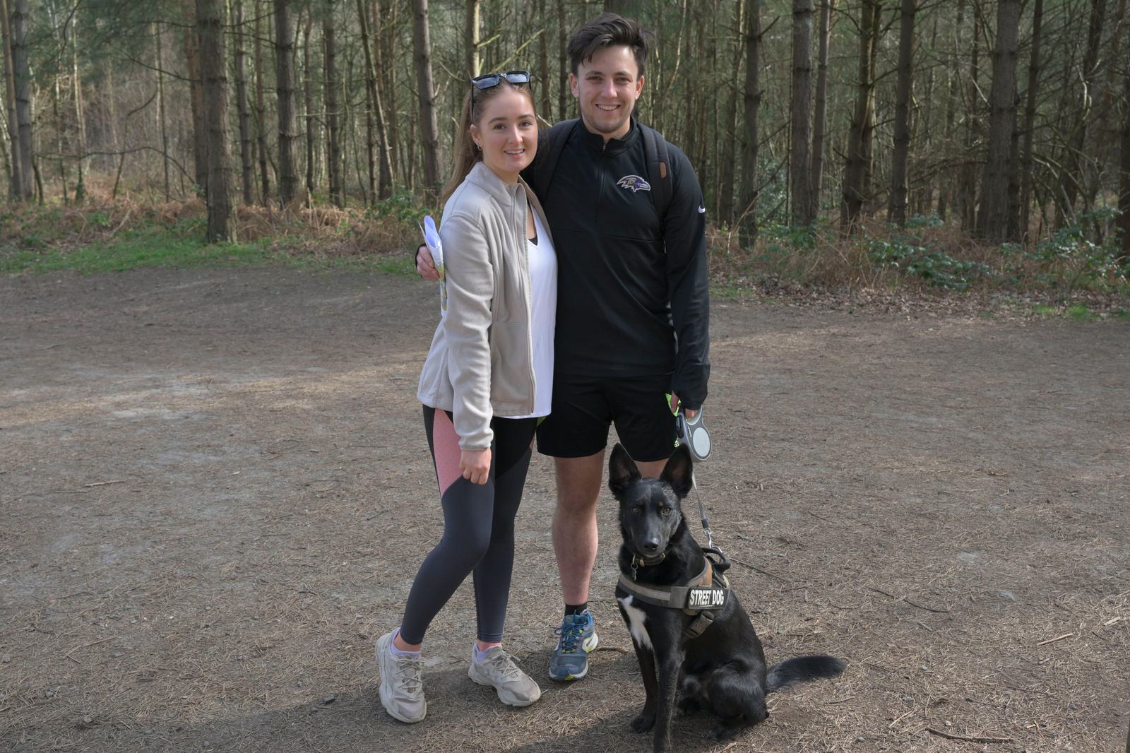 A young woman and man stand together in a wooded area, smiling at the camera. The woman wears a light jacket and leggings, and the man is in athletic wear with a backpack. A black dog with a harness sits in front of them. Trees and dirt ground surround them.