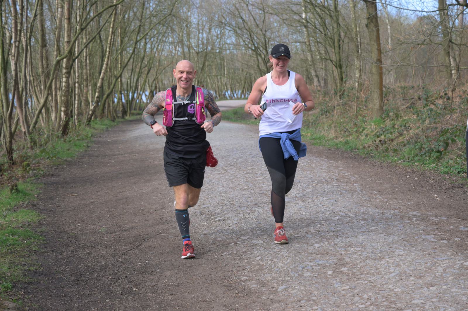 Two runners, a bald tattooed man and a woman in a cap, are running on a gravel path through a forested area. Both are smiling and wearing athletic gear, including shorts, tanks, and running vests. Trees with sparse foliage line the path.