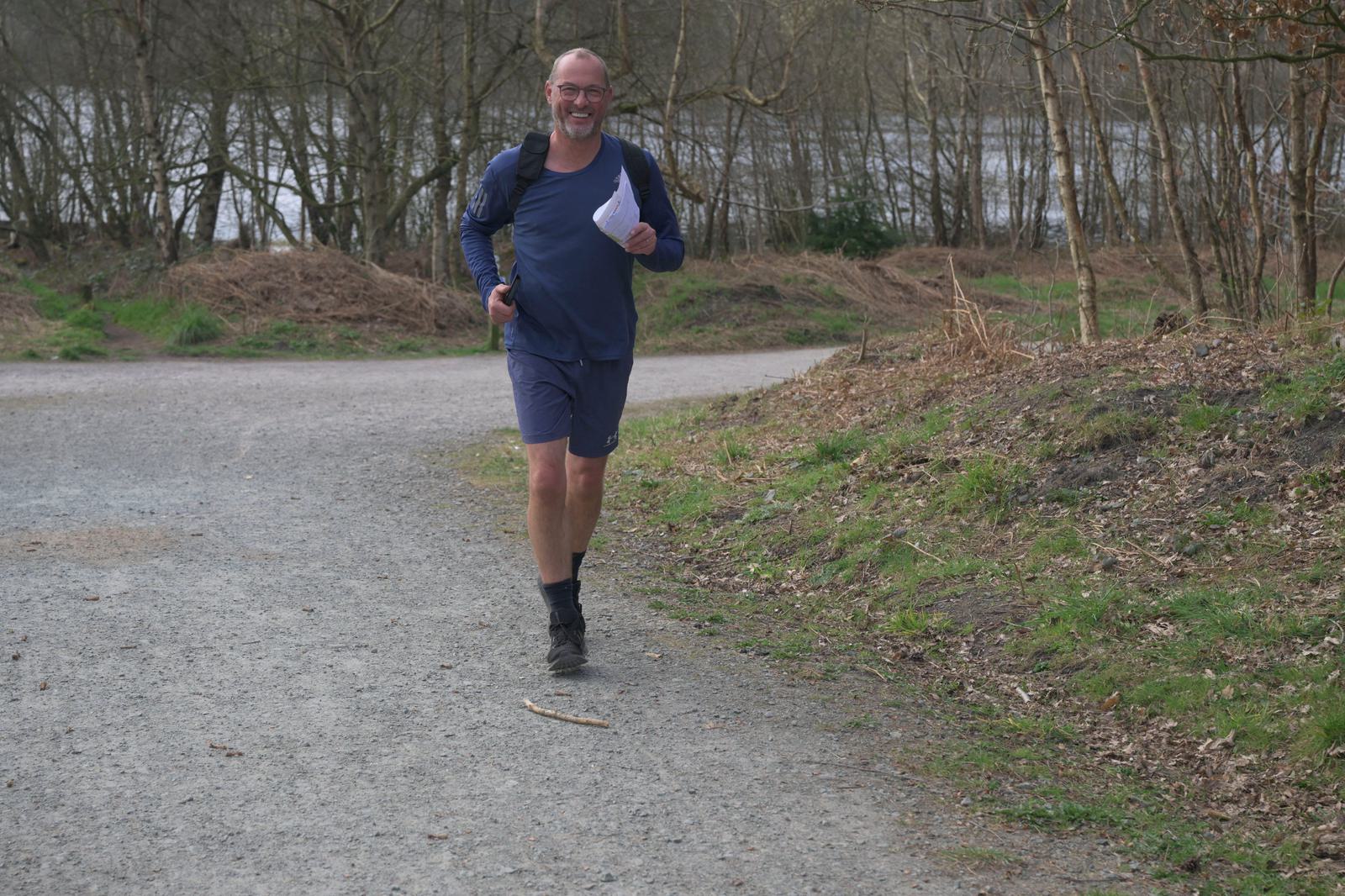 A smiling man with glasses, wearing a navy athletic outfit and black shoes, jogs on a gravel path in a wooded area. He holds a piece of paper in one hand and a jacket draped over his shoulders. Trees and a body of water are visible in the background.