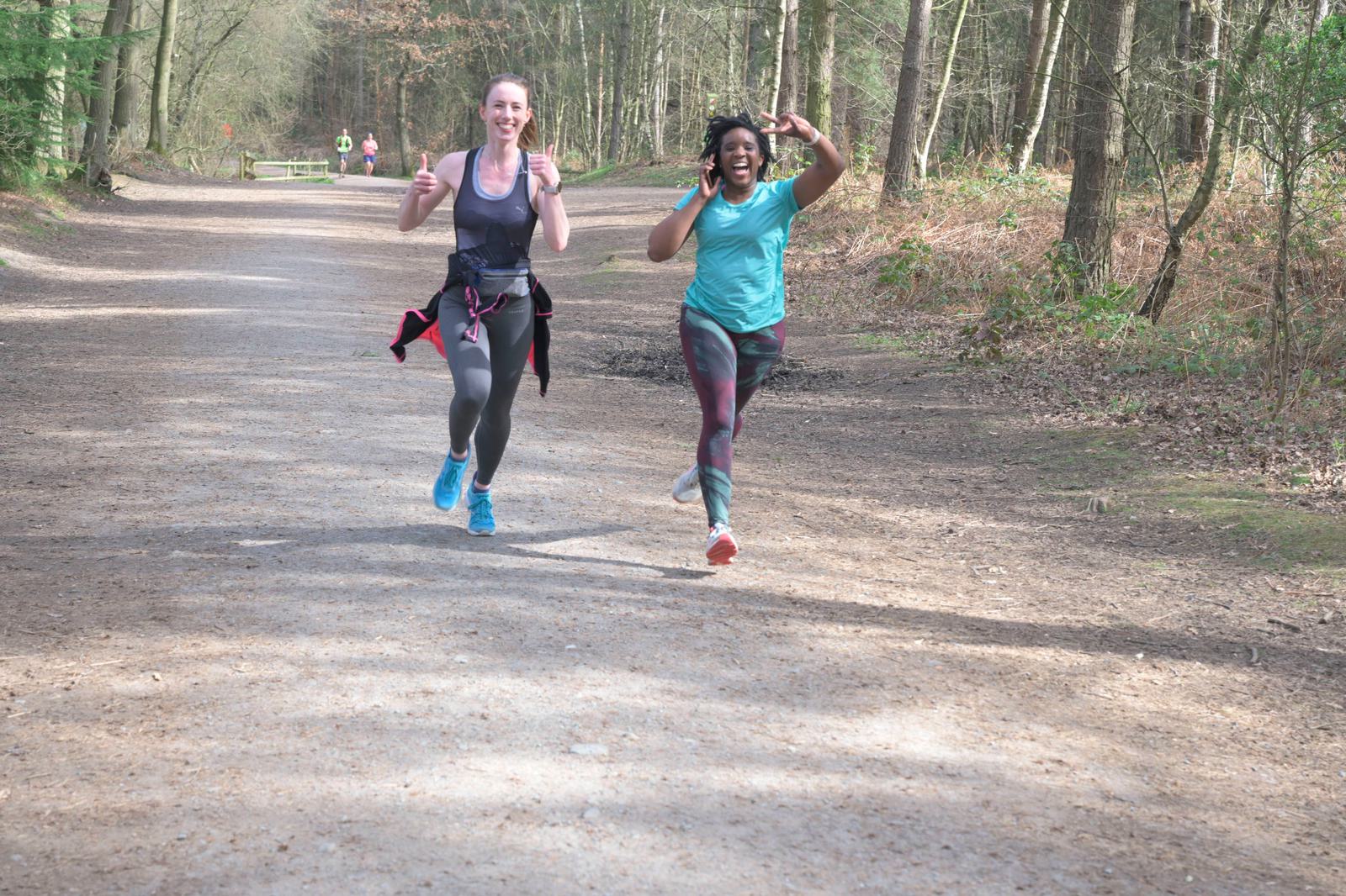 Two women are running joyfully on a woodland trail, smiling and giving thumbs up and peace sign gestures. They are dressed in athletic wear and appear to be enjoying a sunny day of exercise amidst tall trees and natural surroundings.