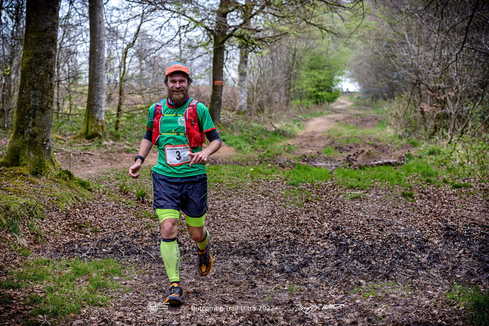 A runner wearing a green shirt, red vest, and yellow leg sleeves runs on a forest trail during a race. Bib number "3" is visible on the front of his shirt. The ground is covered in dry leaves, and trees line both sides of the trail. The sky is partially cloudy.