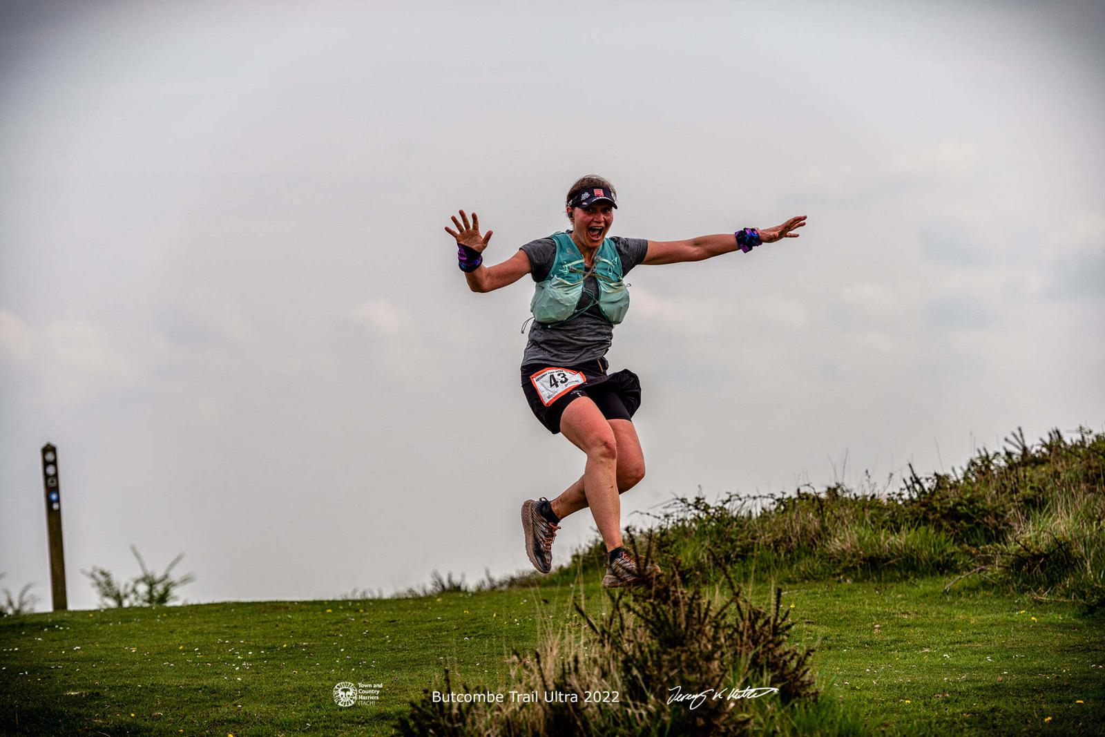 A person wearing running gear and a race bib labeled "180" energetically leaps into the air on a grassy trail. They have a wide smile and raised arms, showcasing their excitement. The scene appears to be during an outdoor trail event, with a cloudy sky in the background.