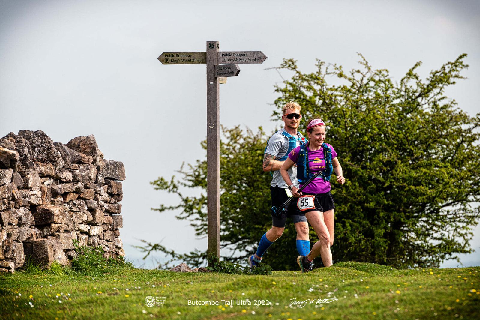 Two trail runners navigate a grassy path, passing a stone wall and a wooden signpost. The woman in the foreground wears a purple shirt and a running vest, while the man behind her stays close. Bright daylight illuminates their path with clear skies in the background.
