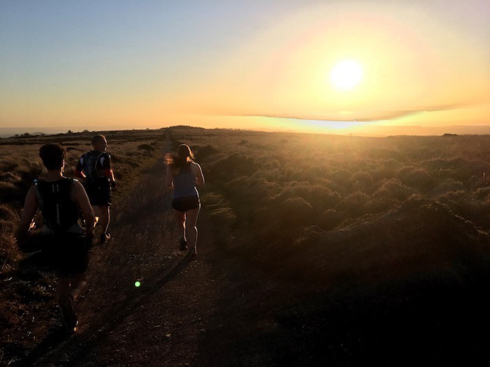 Three people are running on a dirt path through a scenic, dry landscape as the sun sets. The sun is low in the sky, casting a warm, golden glow over the scene. Bushes and uneven terrain surround the trail. The sky is clear, with a few distant clouds on the horizon.