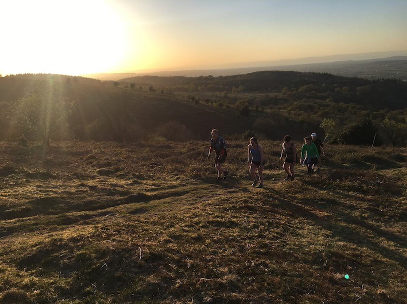 A group of five people hike up a grassy hill at sunset. The sun is low in the sky, casting a golden hue over the landscape. Rolling hills and distant mountains are visible in the background. The path they are on is surrounded by a natural, rugged terrain.
