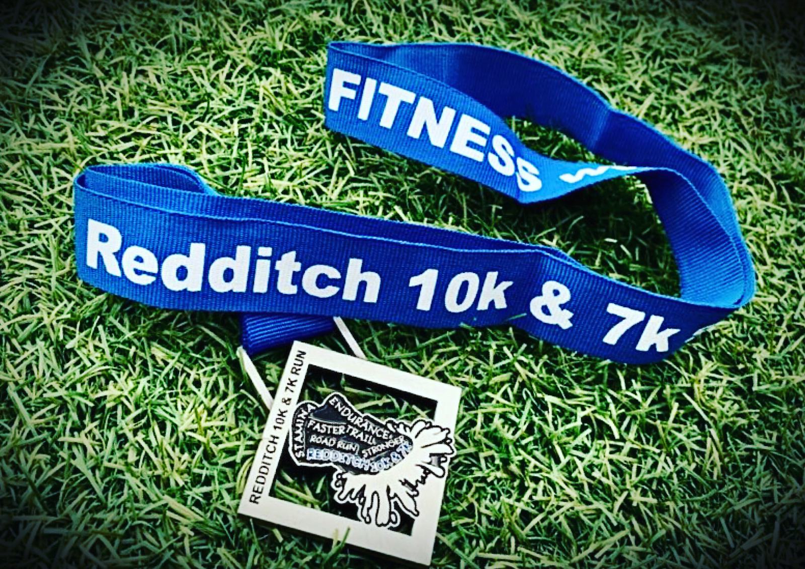 A blue lanyard with the words "FITNESS Redditch 10k & 7k" printed on it. Attached to the lanyard is a silver medal with intricate designs, including a bird in flight and the text "Redditch 10K & 7K Run." The medal and lanyard rest on grass.