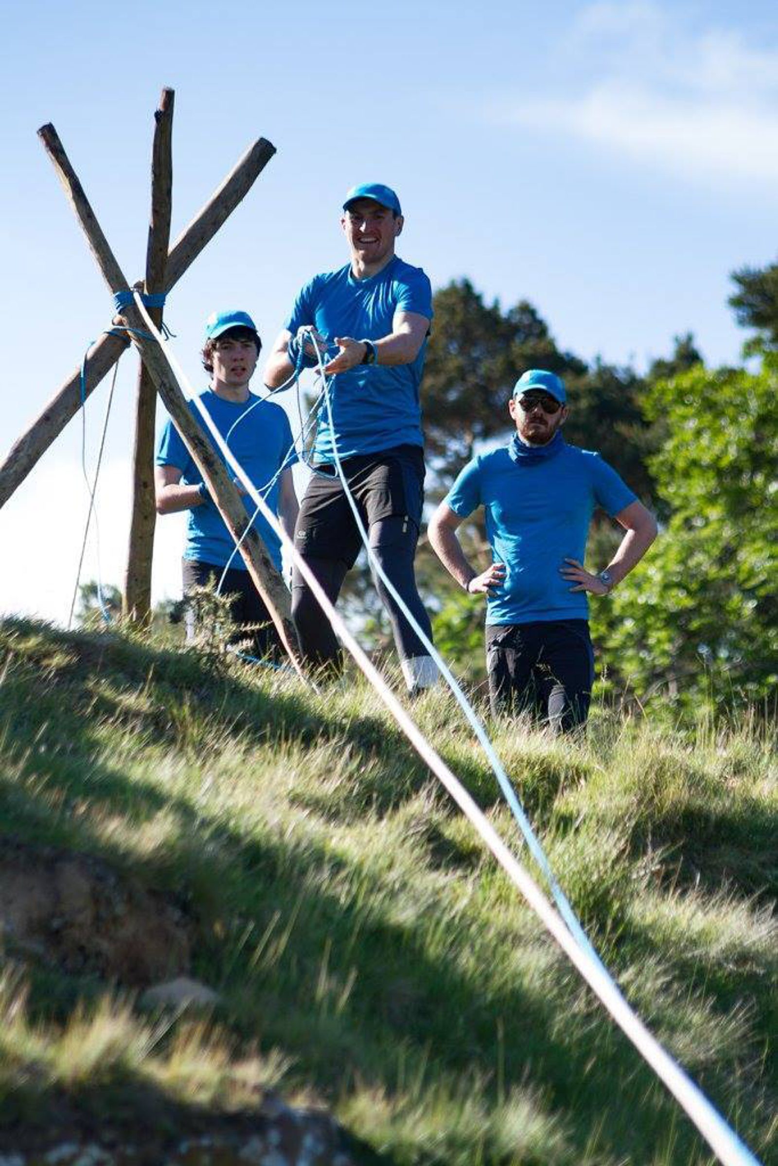 Three men in blue t-shirts and caps are working together on an outdoor project involving ropes and wooden stakes. They are standing on a grassy slope with trees in the background, appearing focused on their task. The weather is sunny and clear.