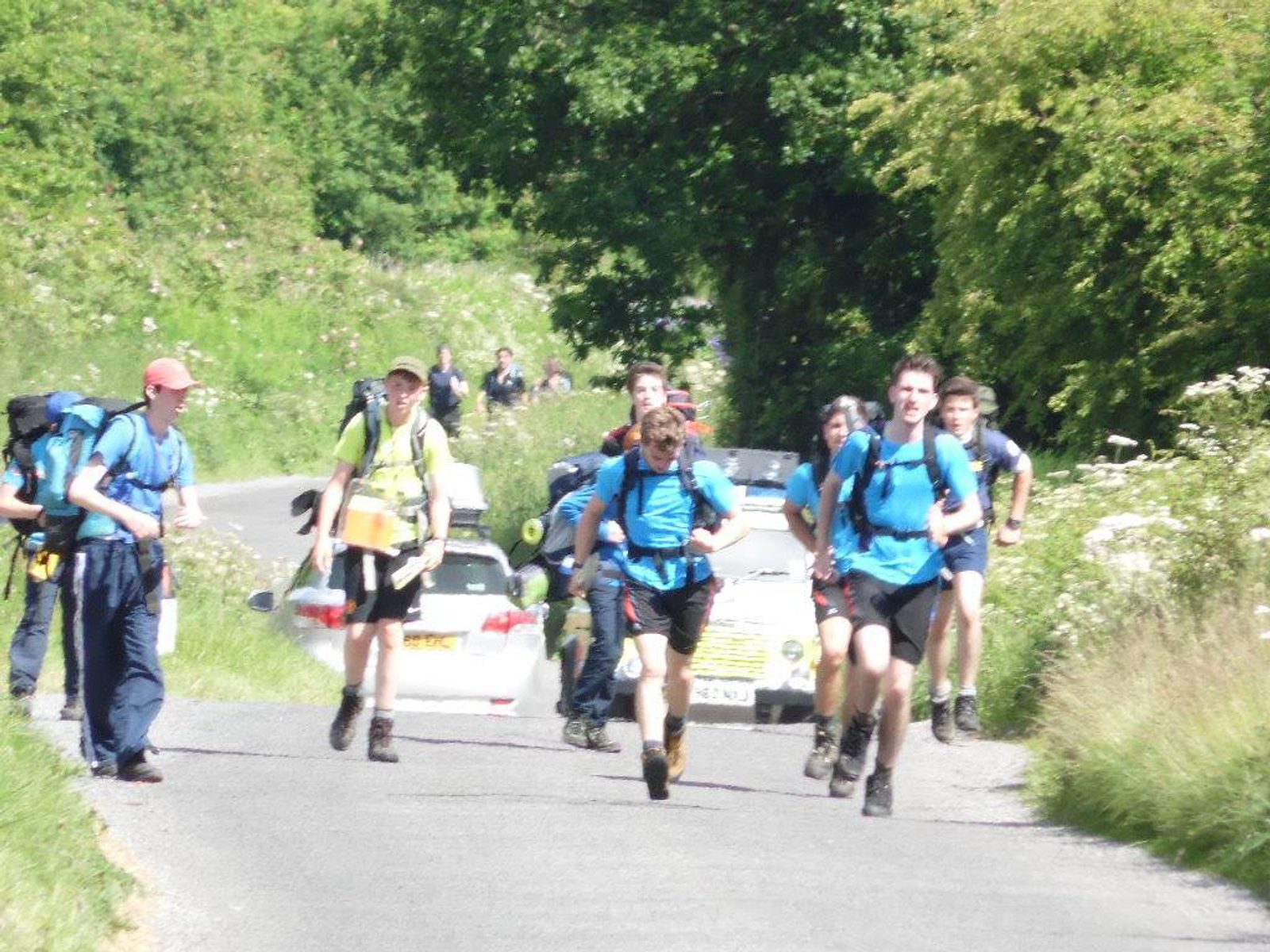 A group of hikers, dressed in blue shirts with backpacks, walk and jog along a country road surrounded by greenery. Several cars are parked in the background, and the hikers appear to be enjoying a sunny day outdoors.