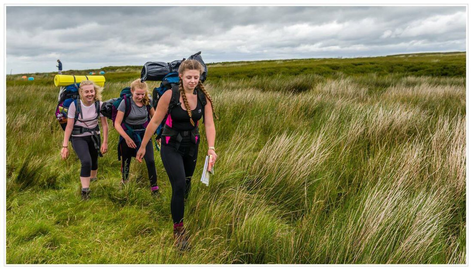 Three people are hiking through tall grass under a cloudy sky. They each carry large backpacks and appear to be navigating with a map. The landscape around them is open and covered with dense grass.