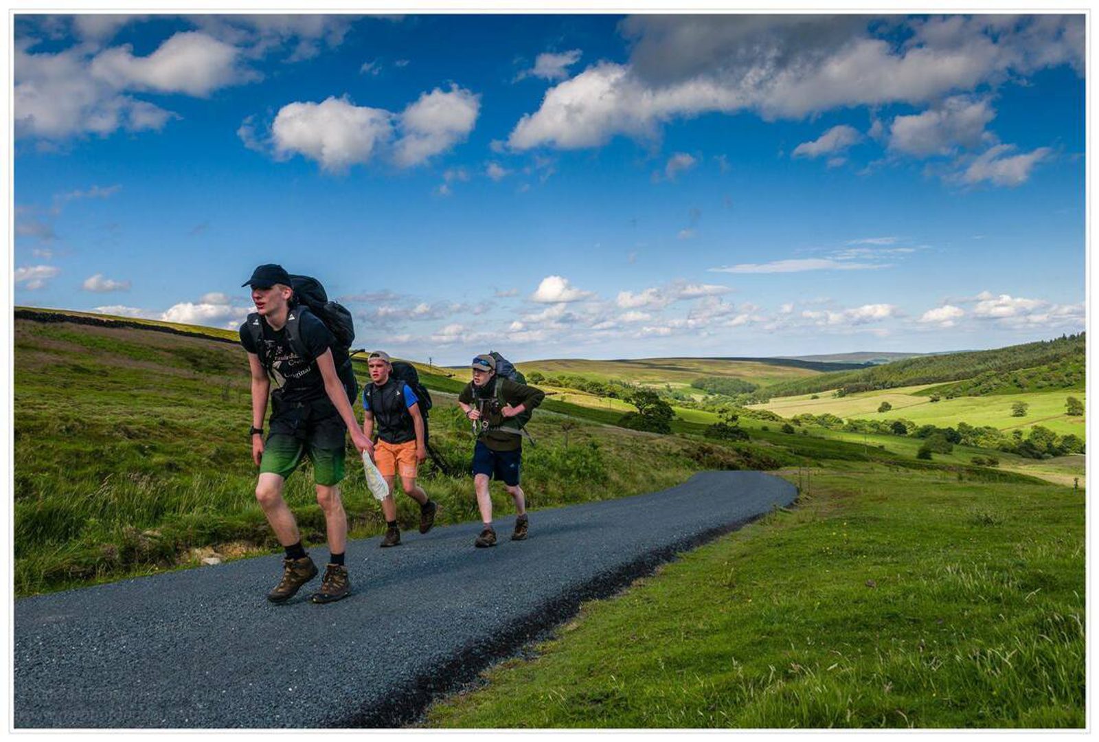 Three hikers with backpacks walk uphill on a paved road through a green landscape under a blue sky with white clouds. The terrain is hilly and expansive, with fields and hills in the background.