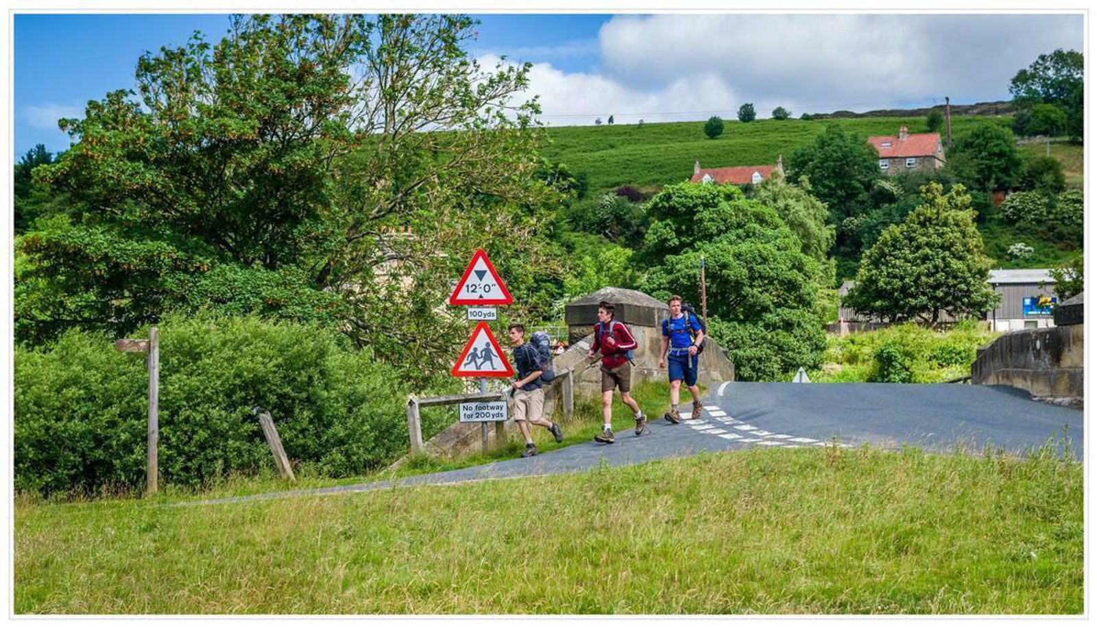 Three people are running on a rural road that curves to the right. There are two road signs indicating a steep 22% incline and the presence of sheep. Lush green fields, trees, and houses with red roofs are in the background under a partly cloudy sky.