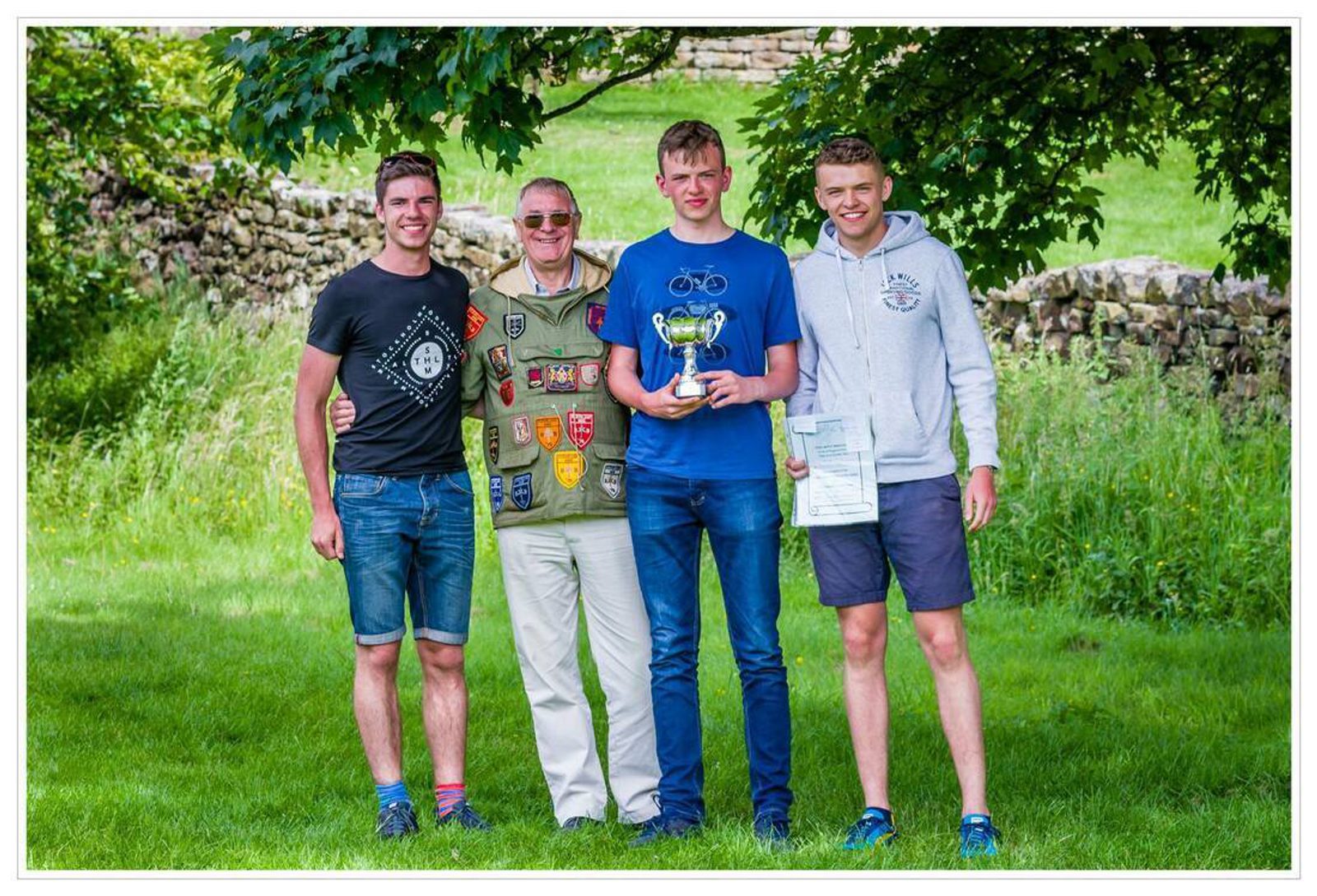 Four men are standing together in a grassy area outdoors, smiling at the camera. One man, wearing a decorated vest, has his hand on the shoulder of another man who is holding a trophy and a certificate. Two other men are standing on either side. Trees and a stone wall are in the background.