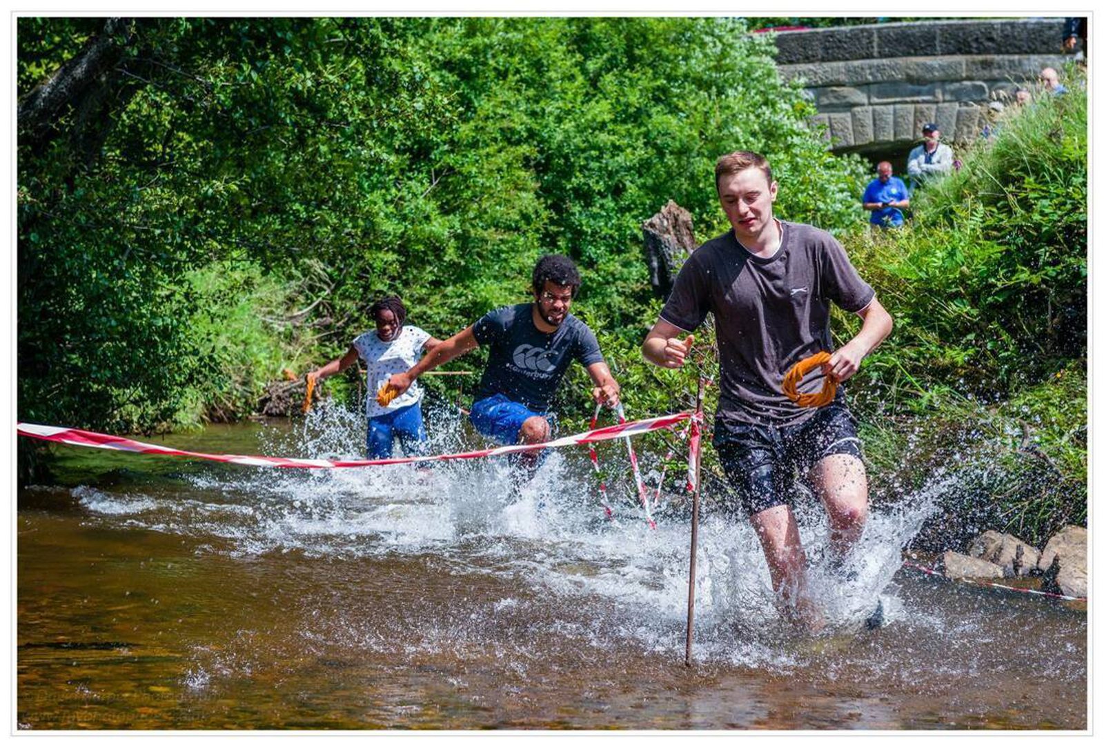 Three people are running through a shallow creek, creating splashes as they go. They appear to be participating in a race, with red and white tape marking the course on either side. The surrounding area is lush with greenery, and a stone bridge is seen in the background.