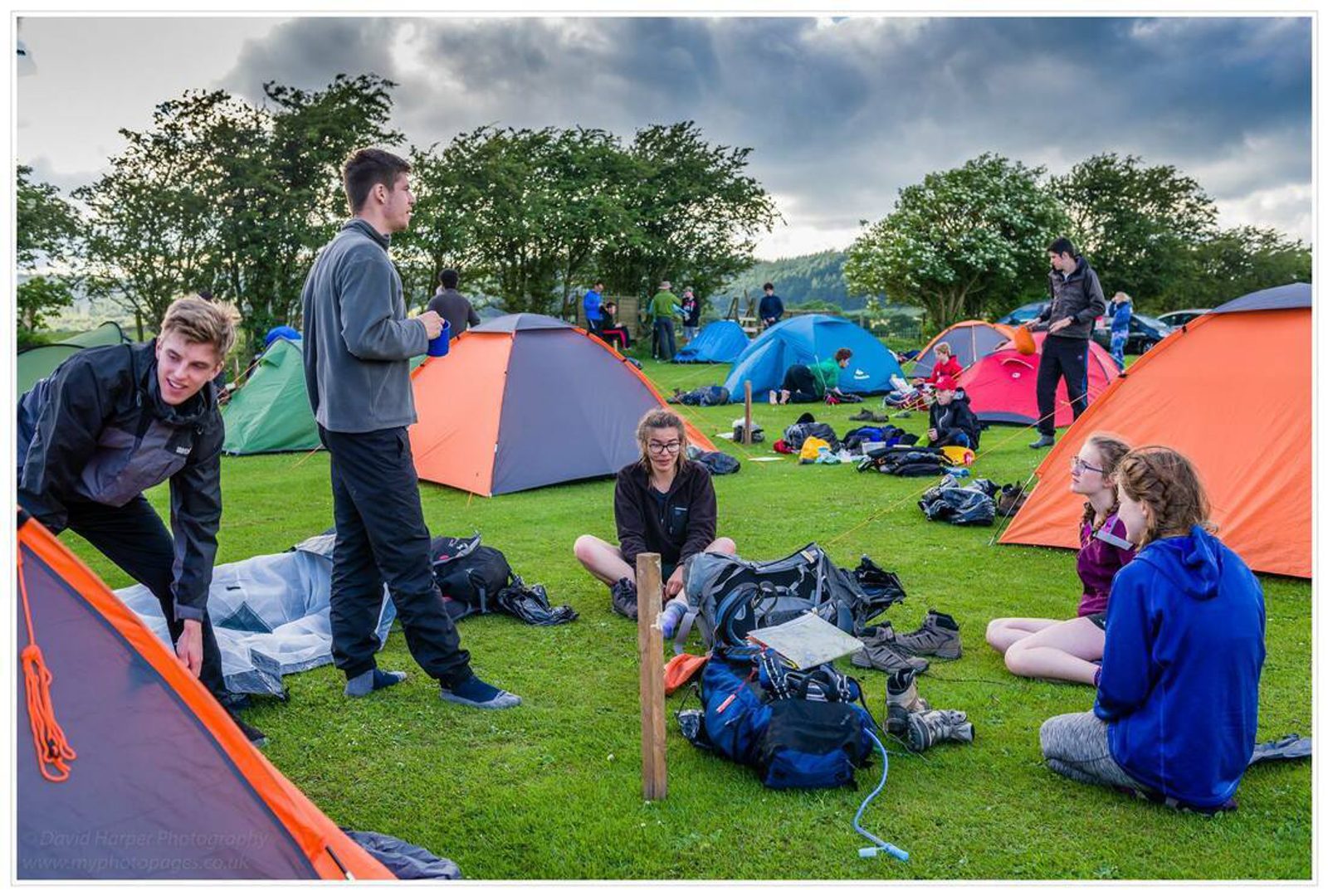 A group of people are gathered on a grassy field with several colorful tents set up around them. They appear to be engaged in various camp activities: setting up tents, organizing gear, and conversing. Trees and a cloudy sky are in the background.