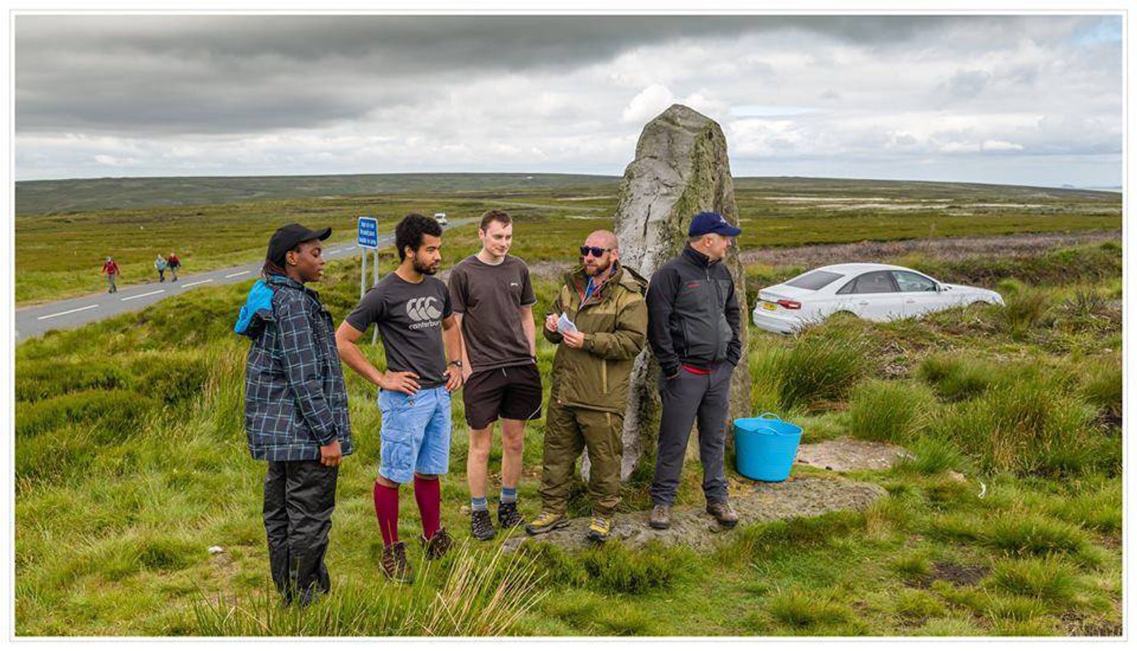 A group of five people stand next to a large stone monument in a grassy, open landscape with a road and a car in the background. Three men wear casual outdoor clothing, while one man is in a rain jacket and another in a cap. The sky is overcast.