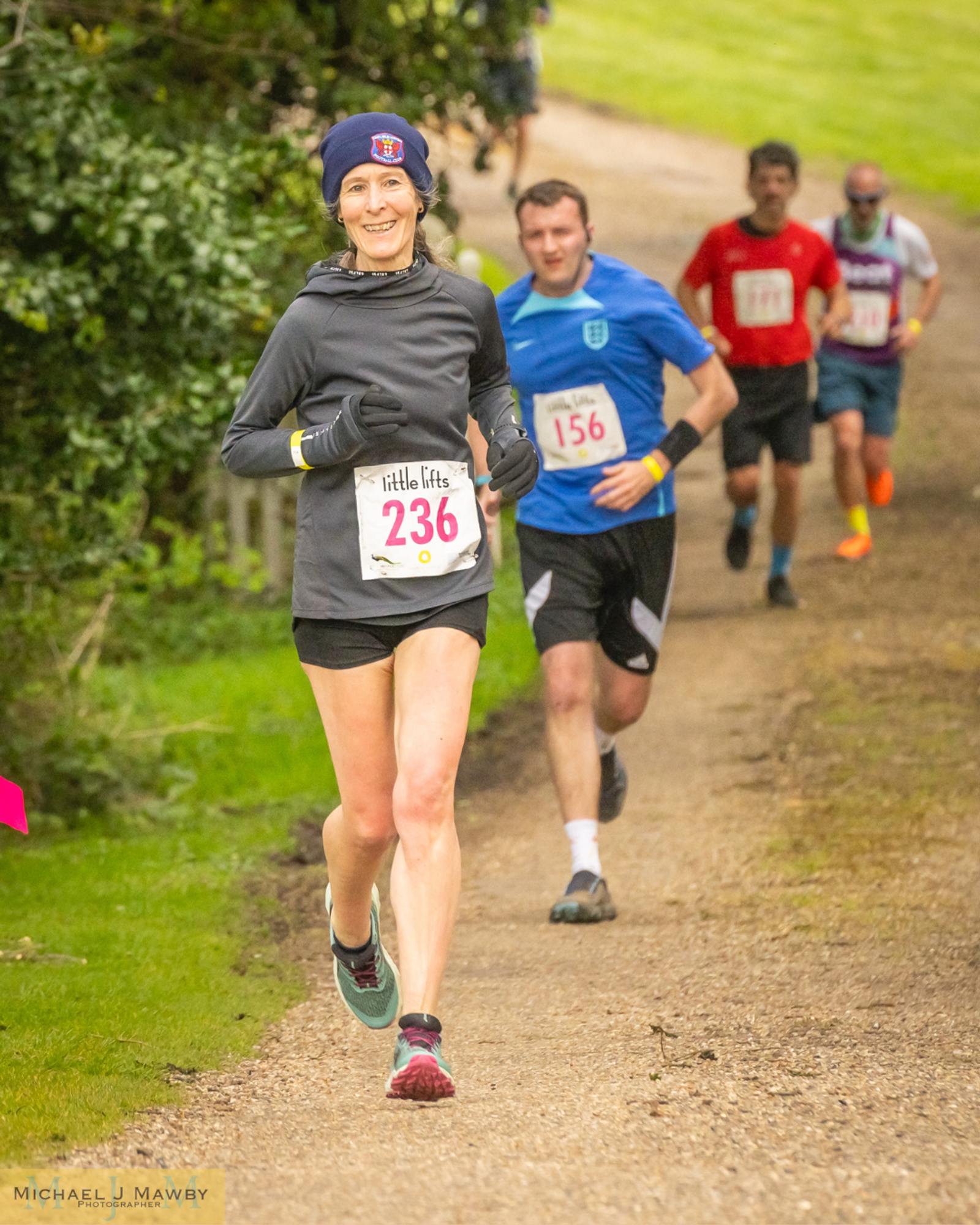 A woman in a black outfit and beanie, wearing the number 236, runs on a gravel path surrounded by greenery. Three men follow her, each in running attire with different colored shirts and race numbers. One is dressed in blue with number 156.