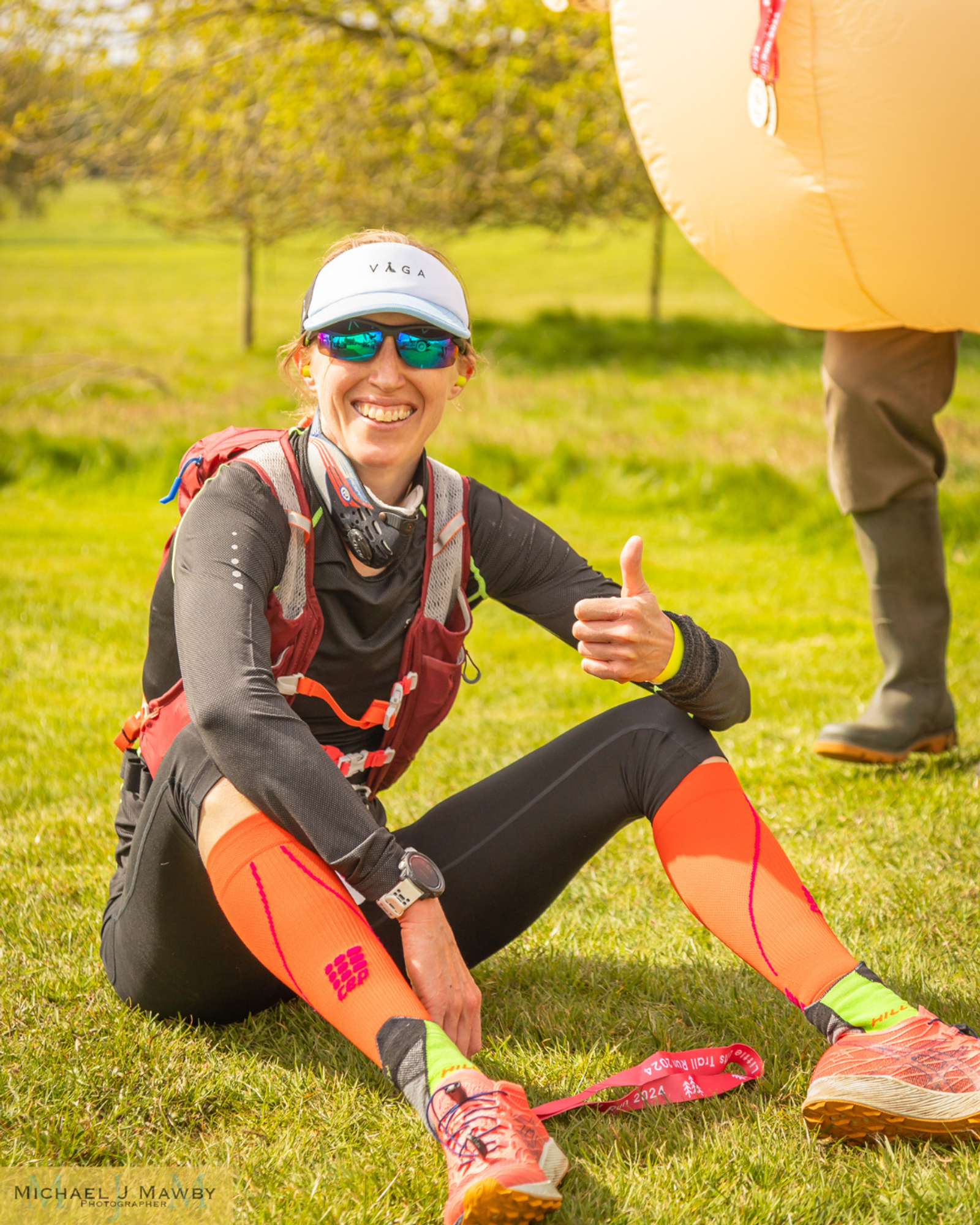 Runner sitting on grass, smiling, wearing a cap, sunglasses, and vibrant athletic wear. She gives a thumbs up, with a medal around her neck. A person with boots and a large prop is partly visible behind.