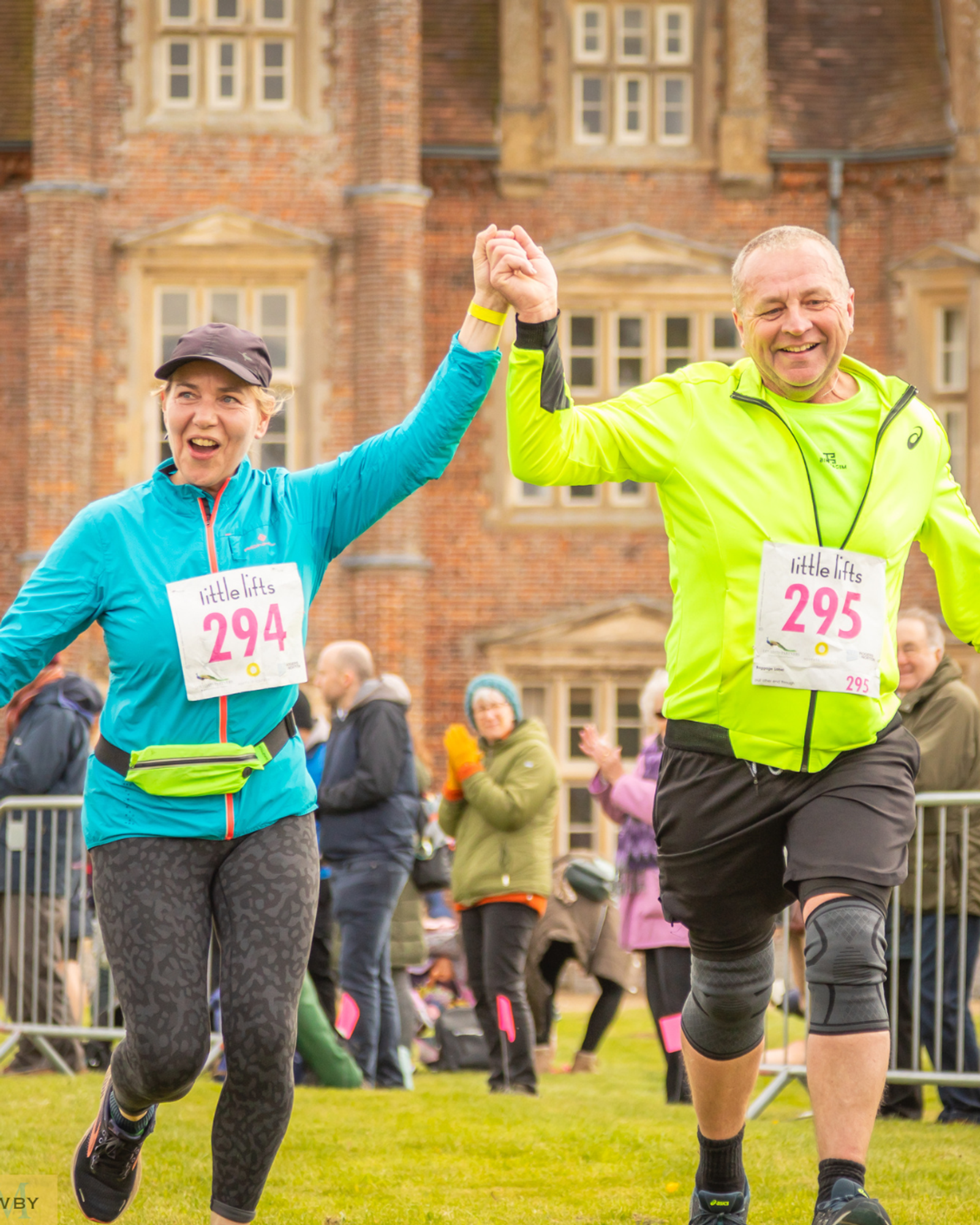 Two joggers, wearing numbered race bibs 294 and 295, smile and hold hands as they cross a grassy finish line. Dressed in bright athletic gear, they celebrate with an enthusiastic crowd and a brick building in the background.