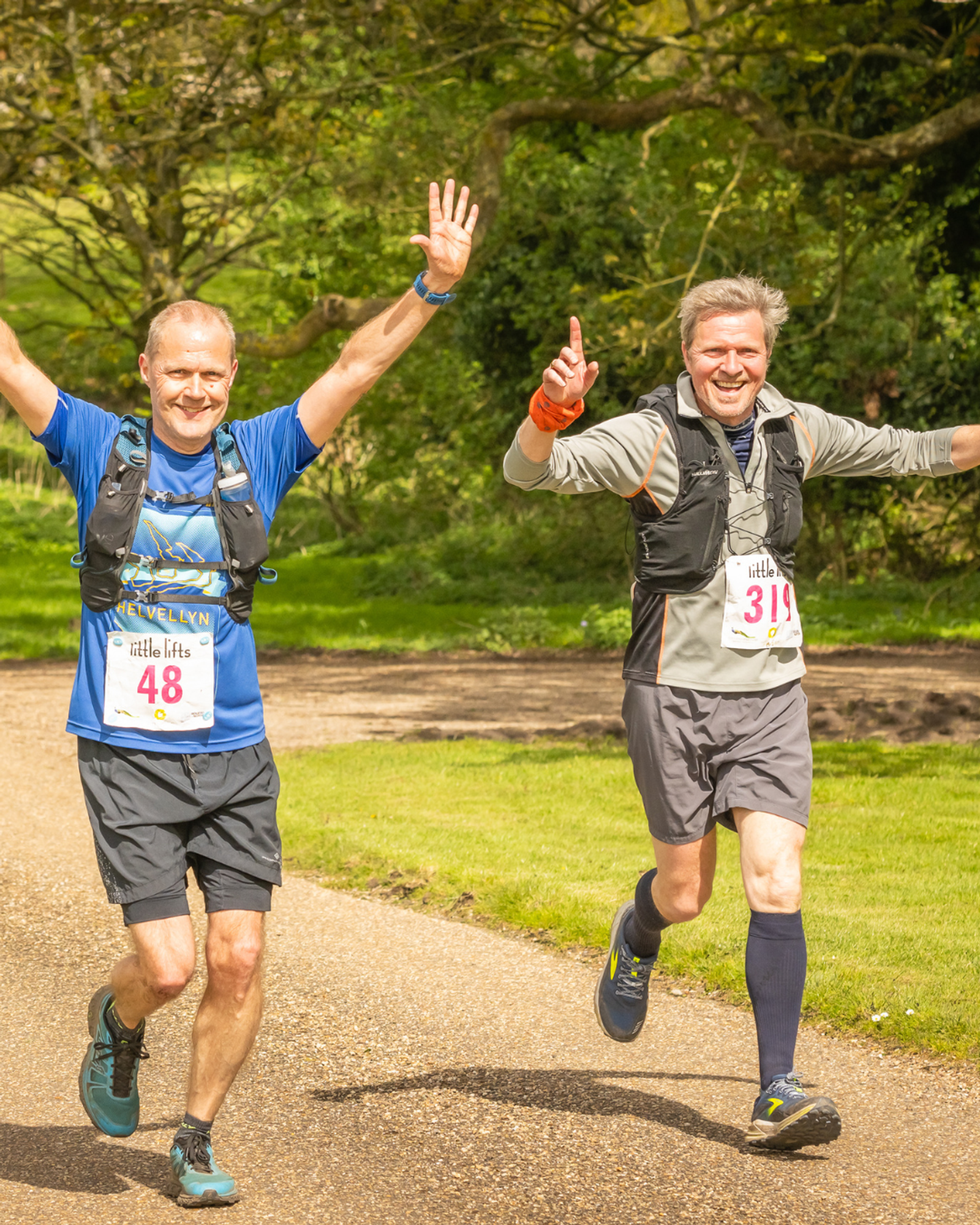 Two men are running on a sunny outdoor path, smiling and raising their arms in celebration. They are wearing running gear and race bibs with numbers 48 and 319. Lush greenery surrounds the path.