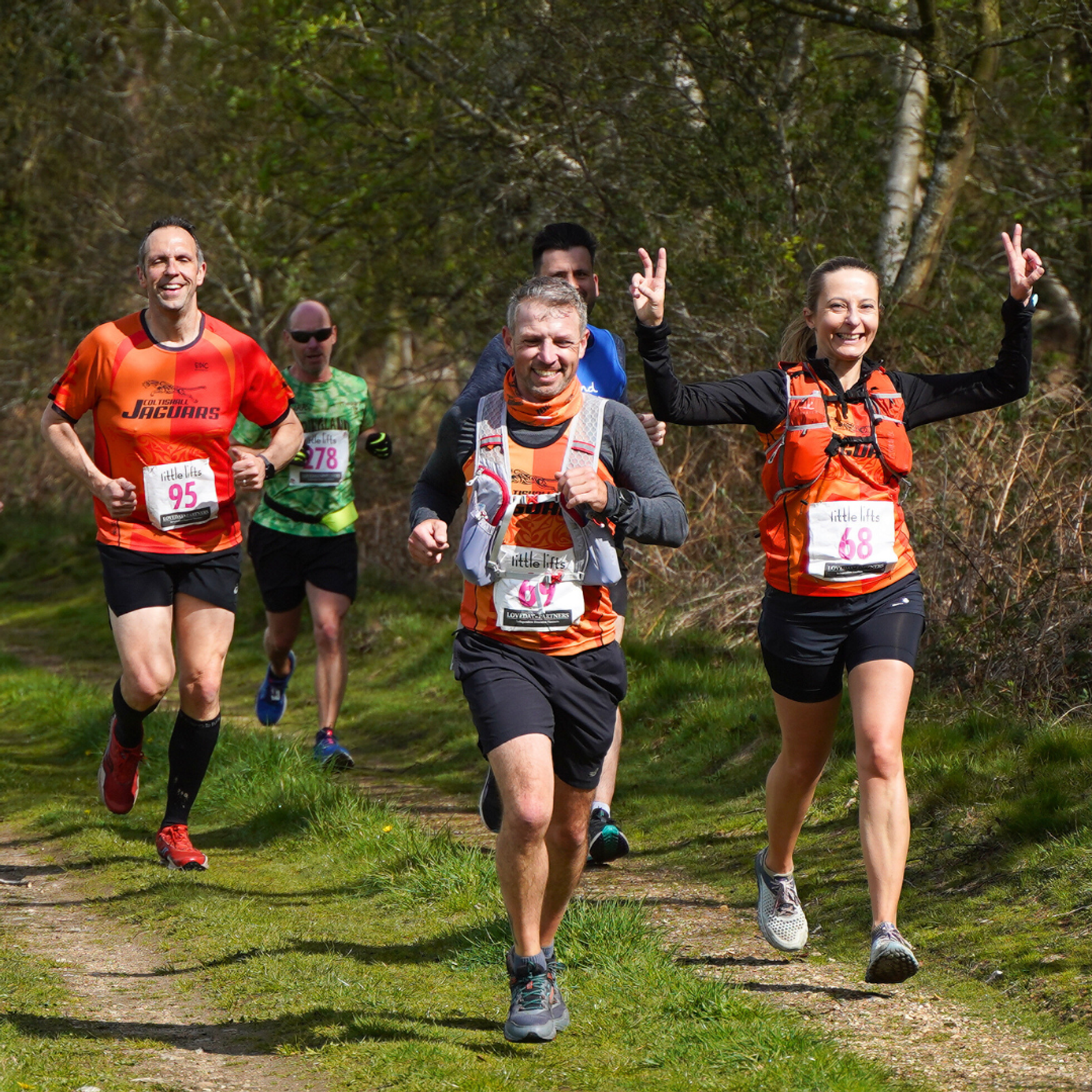 Four people running on a woodland trail during a race. Three of them are wearing orange shirts and race bibs, and one is in green. They are smiling and appear to be enjoying the run, with one person making a peace sign.