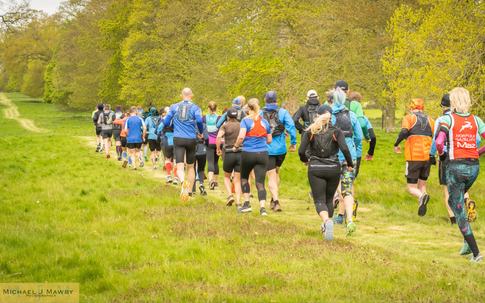 A group of people in athletic gear are running on a grassy trail through a park with lush green trees on either side. The sky is overcast, and the runners are heading away from the camera.