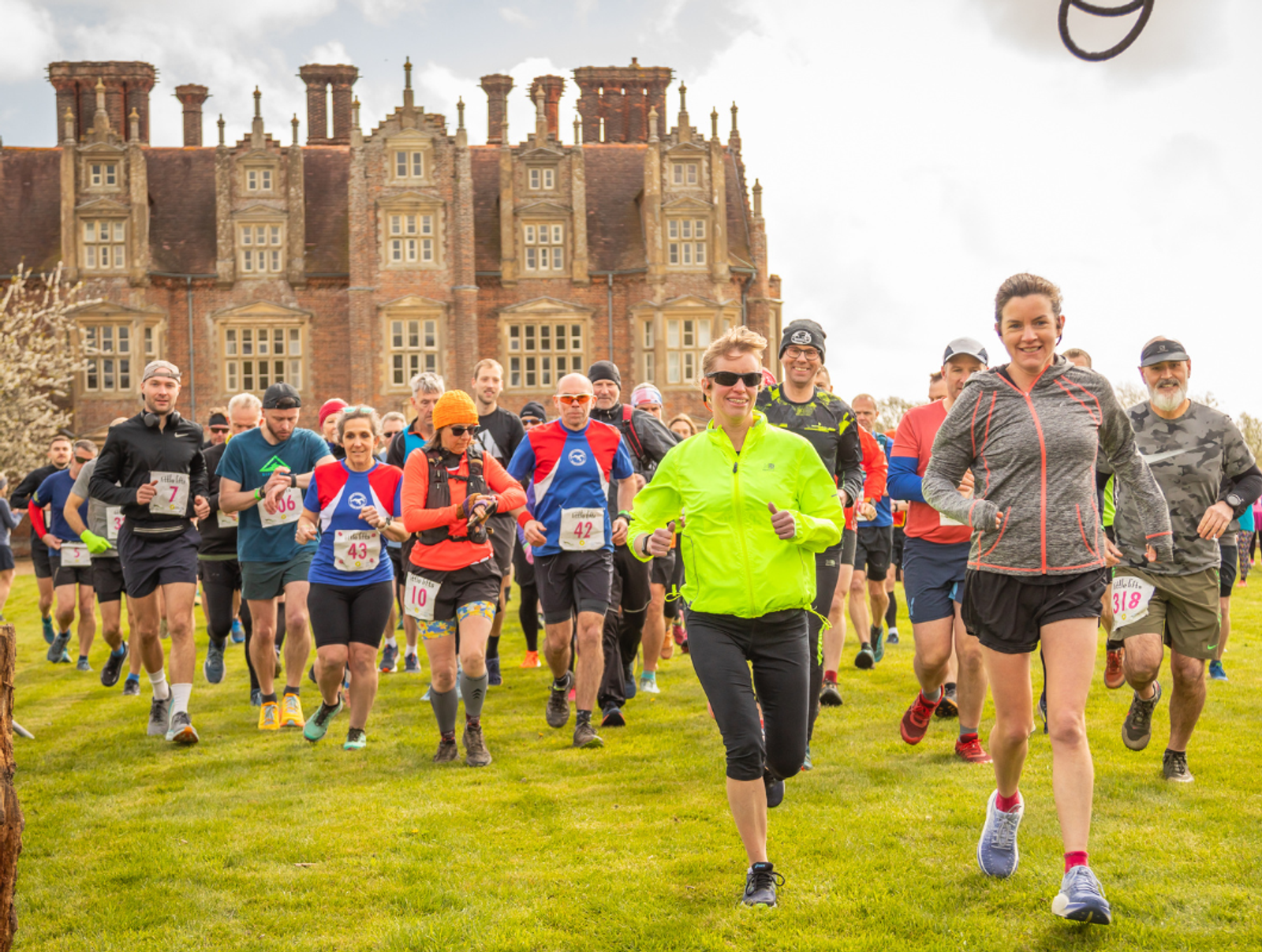 A group of runners in colorful athletic gear participate in a race on a grassy field. A large historic building with ornate architecture is in the background. The runners are smiling as they move forward.