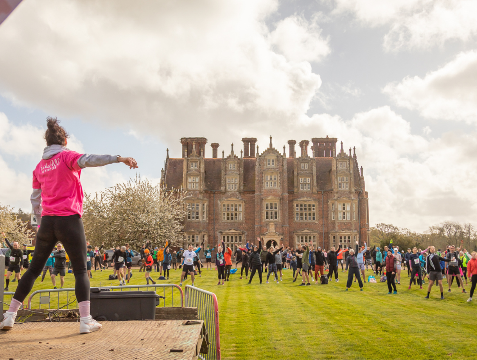 A group of people exercise on a large green lawn in front of a historic mansion. A person stands on a platform in the foreground, leading the workout. The sky is cloudy, and trees with blossoms line the lawn.