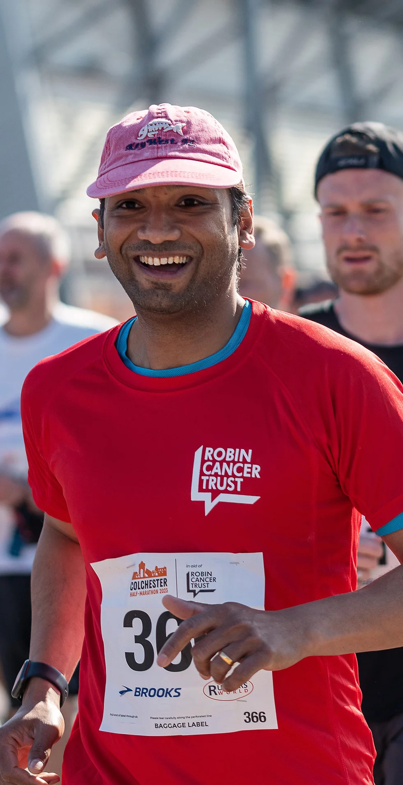 A smiling runner wearing a red Robin Cancer Trust shirt and a pink cap is participating in a race. Other runners can be seen in the background. The runner is holding a timing device or tracker in hand and a race number 366 is visible on his shirt.