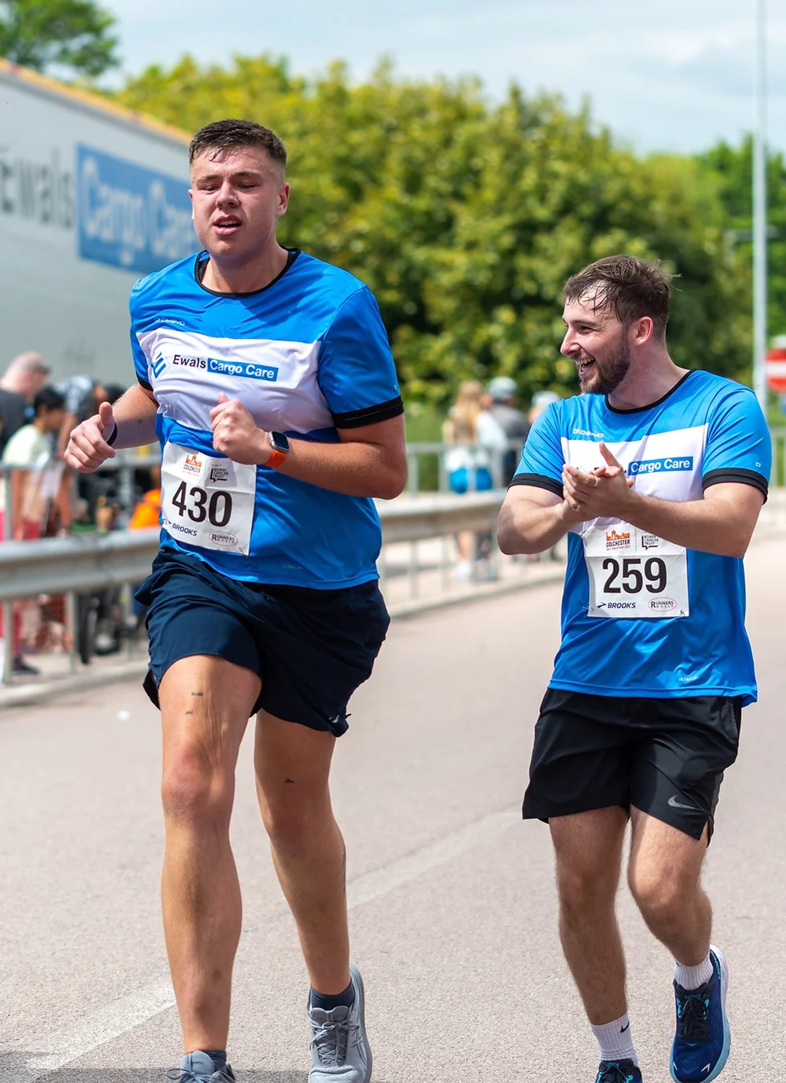 Two men are participating in a marathon, both wearing blue athletic shirts. The man on the left, bib number 430, is running ahead, while the man on the right, bib number 259, is encouraging him with applause. A crowd and greenery are visible in the background.