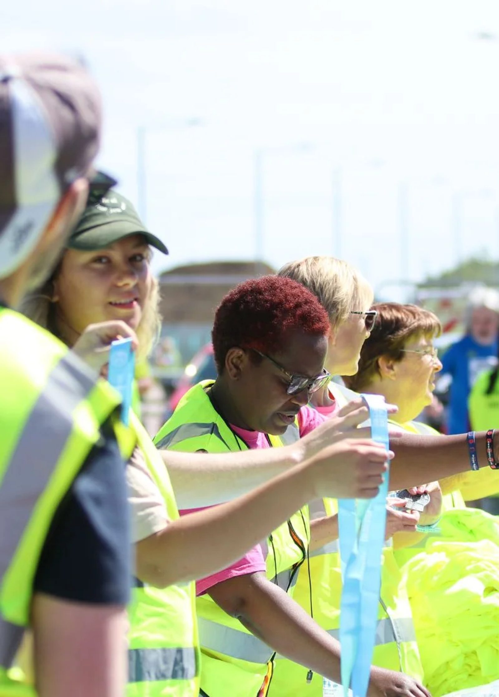 A group of volunteers wearing bright yellow vests distribute what seem to be tickets or ribbons at an outdoor event. They are smiling and engaged, with a diverse mix of individuals participating in the activity.