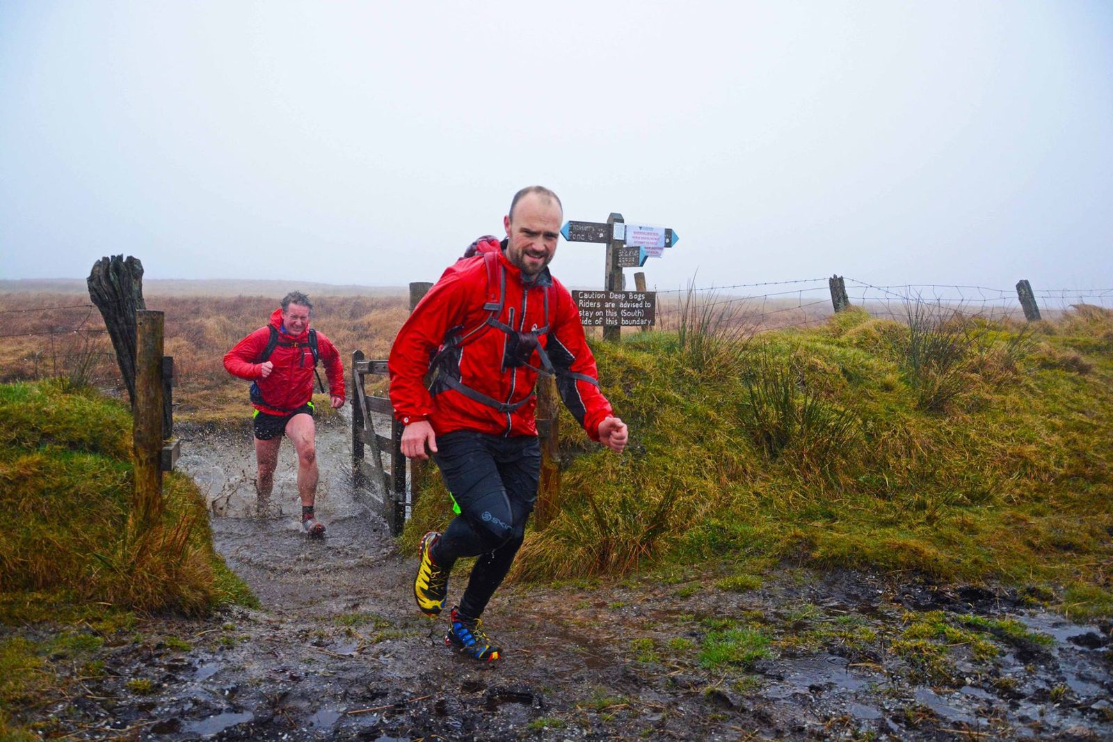 Two people in red jackets run along a muddy path in a foggy, rural landscape. One jumps over a puddle, while the other follows. A wooden fence and direction sign are visible in the background. Grass and moorland surround the scene.