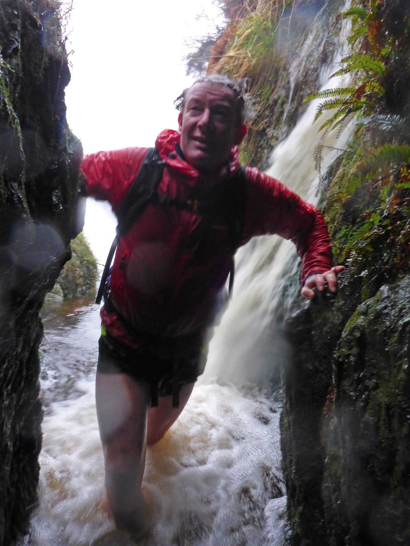 A person in a red jacket stands in a narrow rocky gap with water rushing around their legs. They appear to be outdoors, near a waterfall or fast-flowing stream, against a backdrop of wet rocks and ferns.