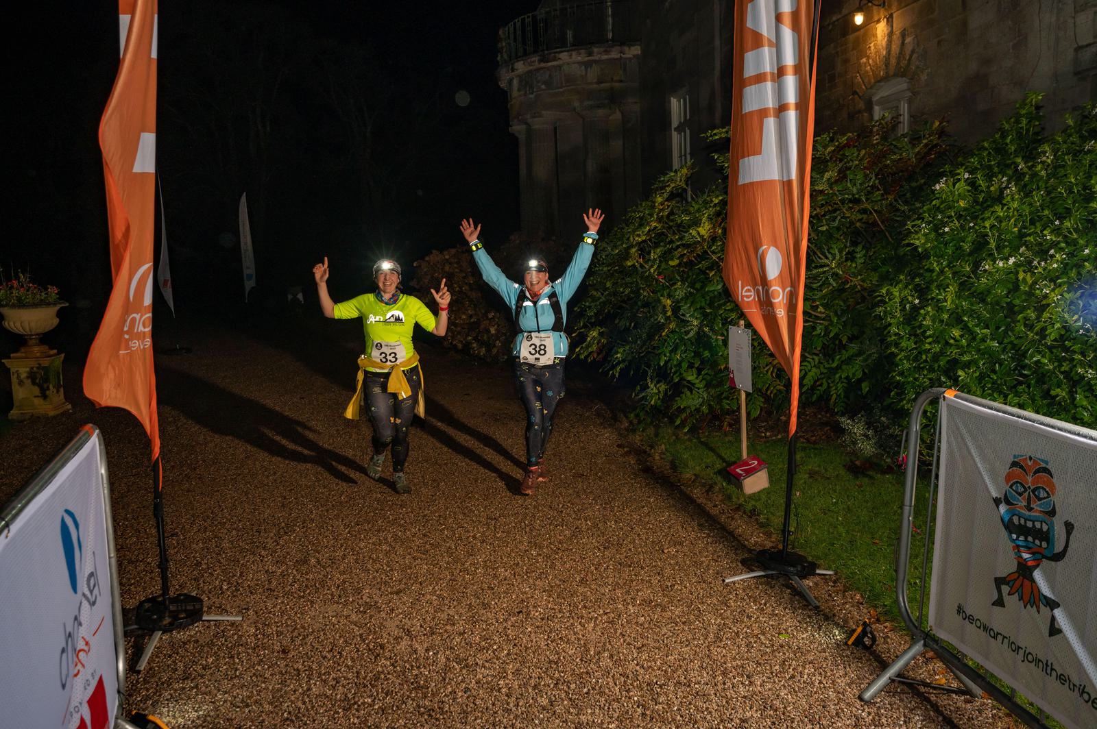 Two nighttime runners wearing headlamps and numbered bibs, 33 and 38, celebrate as they approach the finish line on a gravel path at an event. Brightly colored flags and banners mark the finish area, and an ornate building can be seen in the background.