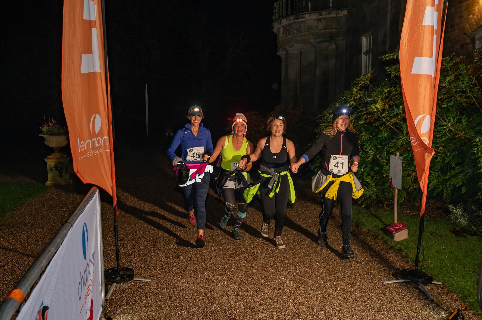 Four women are running at night, smiling and holding hands. They wear athletic clothing, headlamps, and race numbers (41, 43, 45, 47). They are approaching a finish line marked by orange banners with "Lennon" branding. A stone building and greenery are visible in the background.