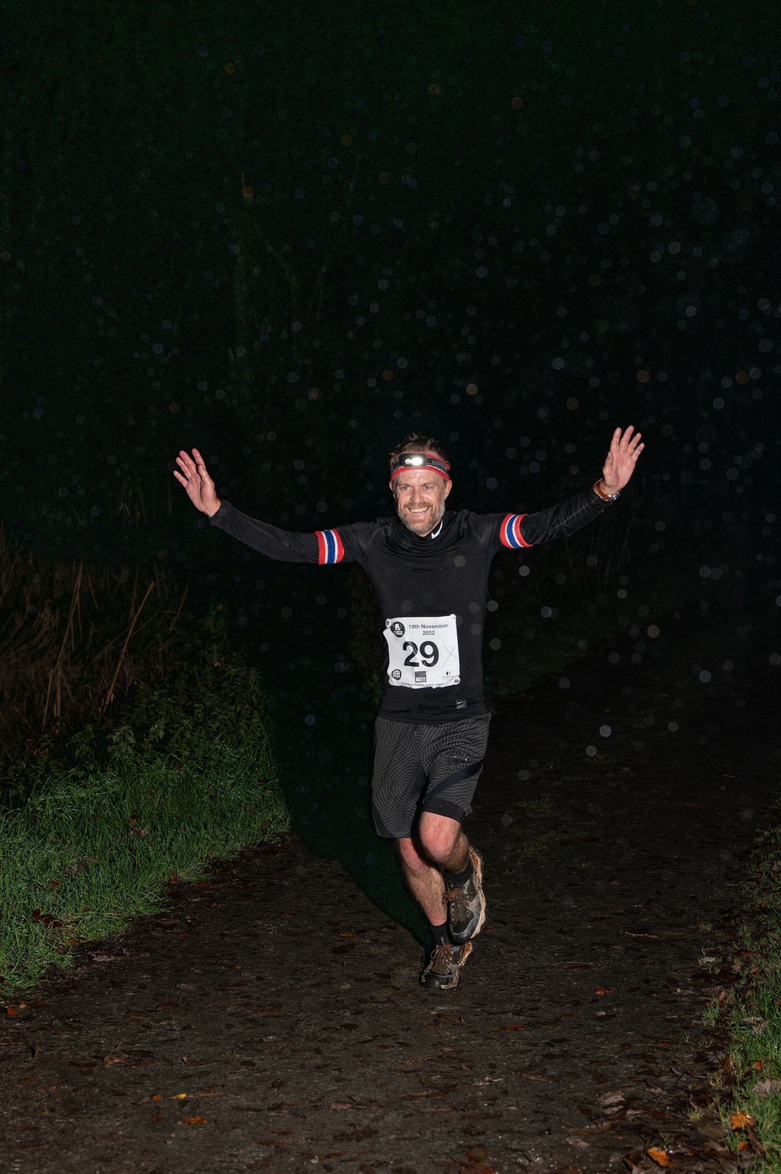 A man running at night on a dimly lit path with his arms raised in victory. He is wearing a numbered race bib, headlamp, black athletic gear, and a smile. The background is dark with faint trees and scattered raindrops reflecting the light.