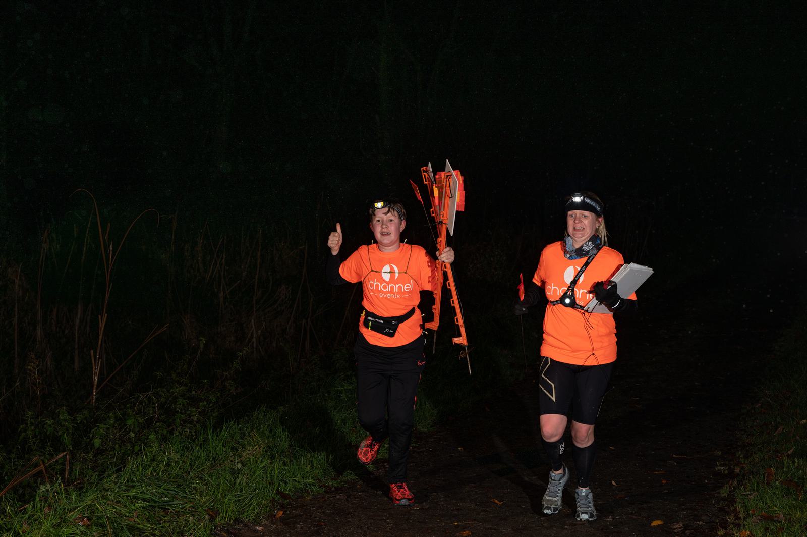 Two runners wearing orange shirts and headlamps jog through a dark wooded area. One holds a signpost, and the other carries a clipboard. They appear determined and energized, possibly participating in a night race or outdoor event.