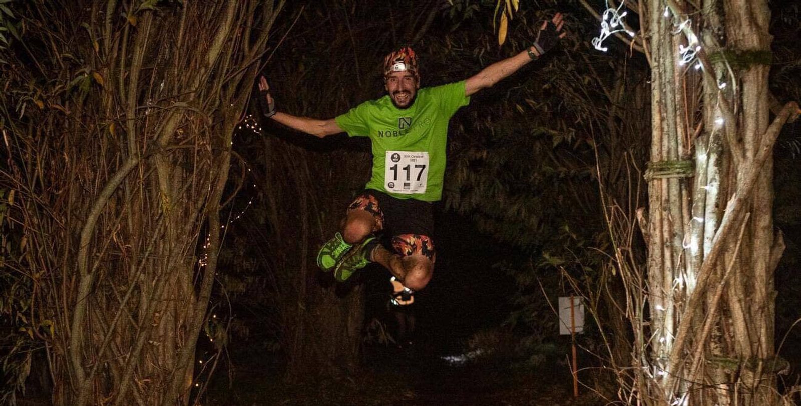 Runner in a green shirt jumps energetically along a dark, wooded trail during the Moonlit Meadow Run 2021. The path is illuminated by soft lights, creating a dynamic and adventurous atmosphere.