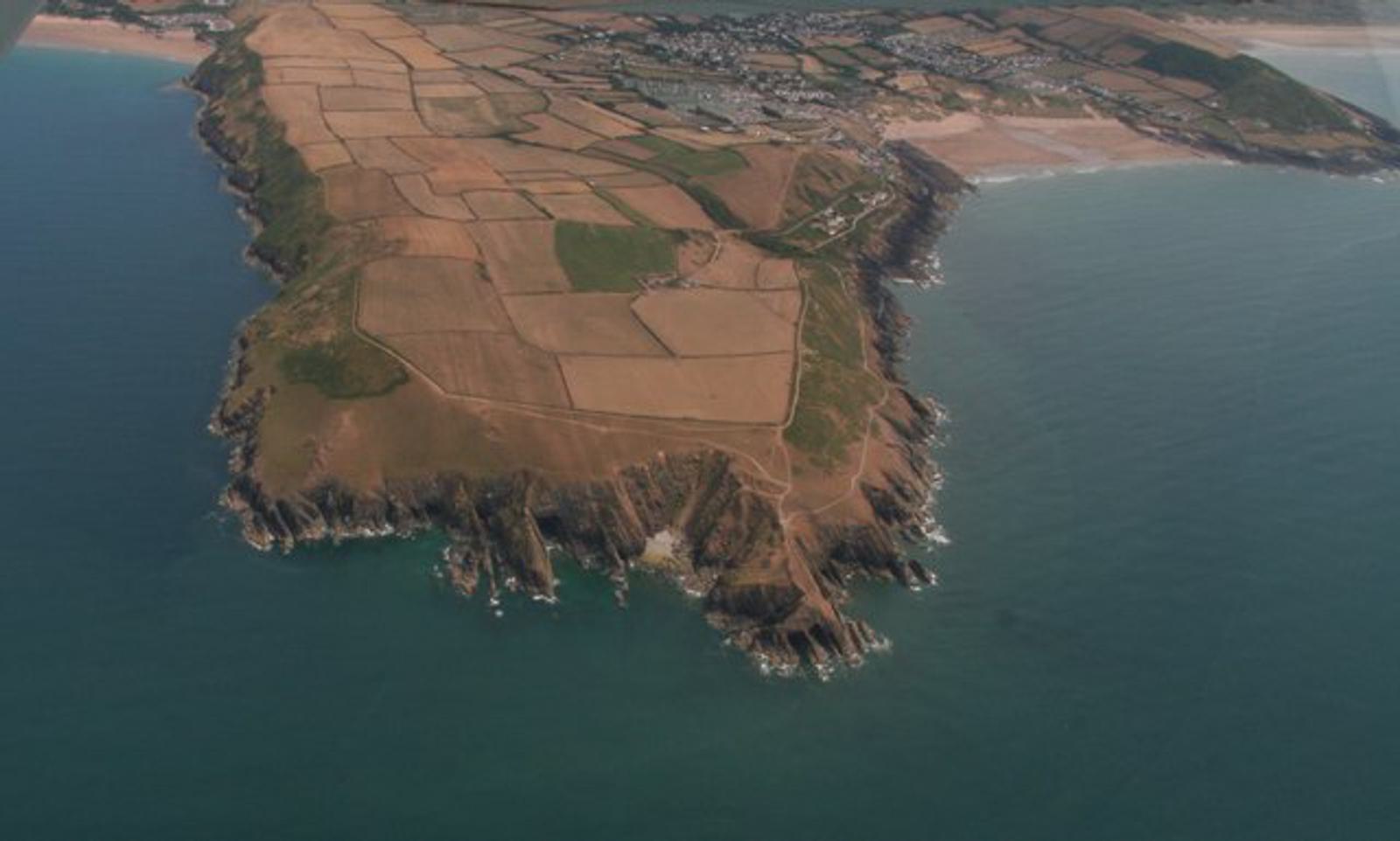 Aerial view of an extensive coastal landscape featuring a rocky peninsula with patchwork fields. The land is surrounded by calm blue ocean waters. Houses and roads are visible in the background near the shoreline.