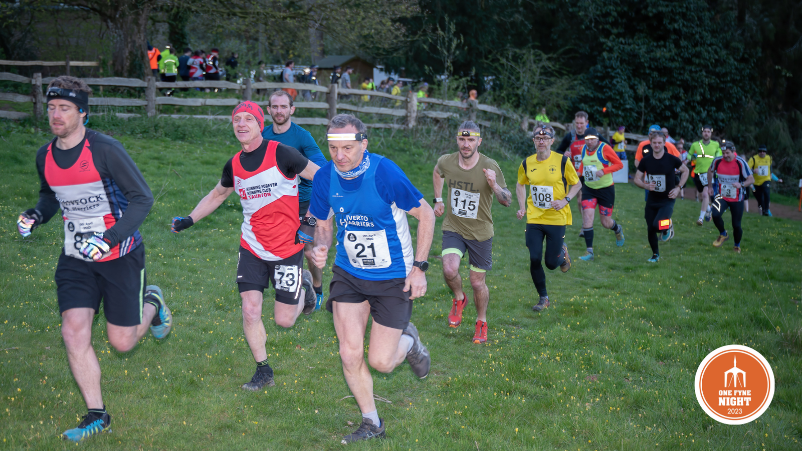 A group of runners, dressed in various athletic gear, race along a grassy path during an organized event. Some wear bibs with numbers while others sport headbands and hats. In the background, spectators and additional runners are visible. A logo for a 2023 night event is at the bottom right.