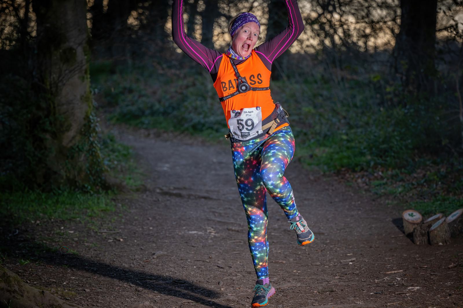 A person wearing a colorful outfit and runner bib number 59 is joyfully jumping on a forested trail. They have on a pink and purple headband, an orange top, and rainbow-colored leggings. The background includes trees and a dirt path.