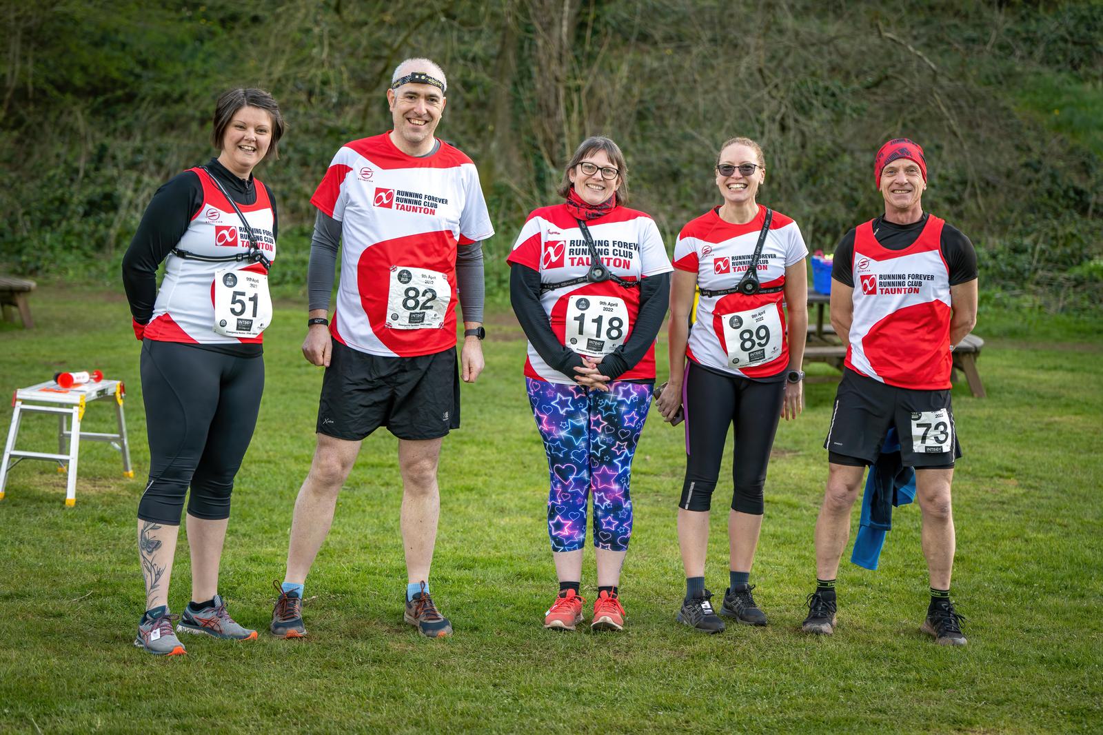 Five runners standing outdoors on grass, posing for a group photo. They are wearing red and white running shirts with various bib numbers. Trees and a pathway are visible in the background. The runners are smiling and appear to be participating in a race event.