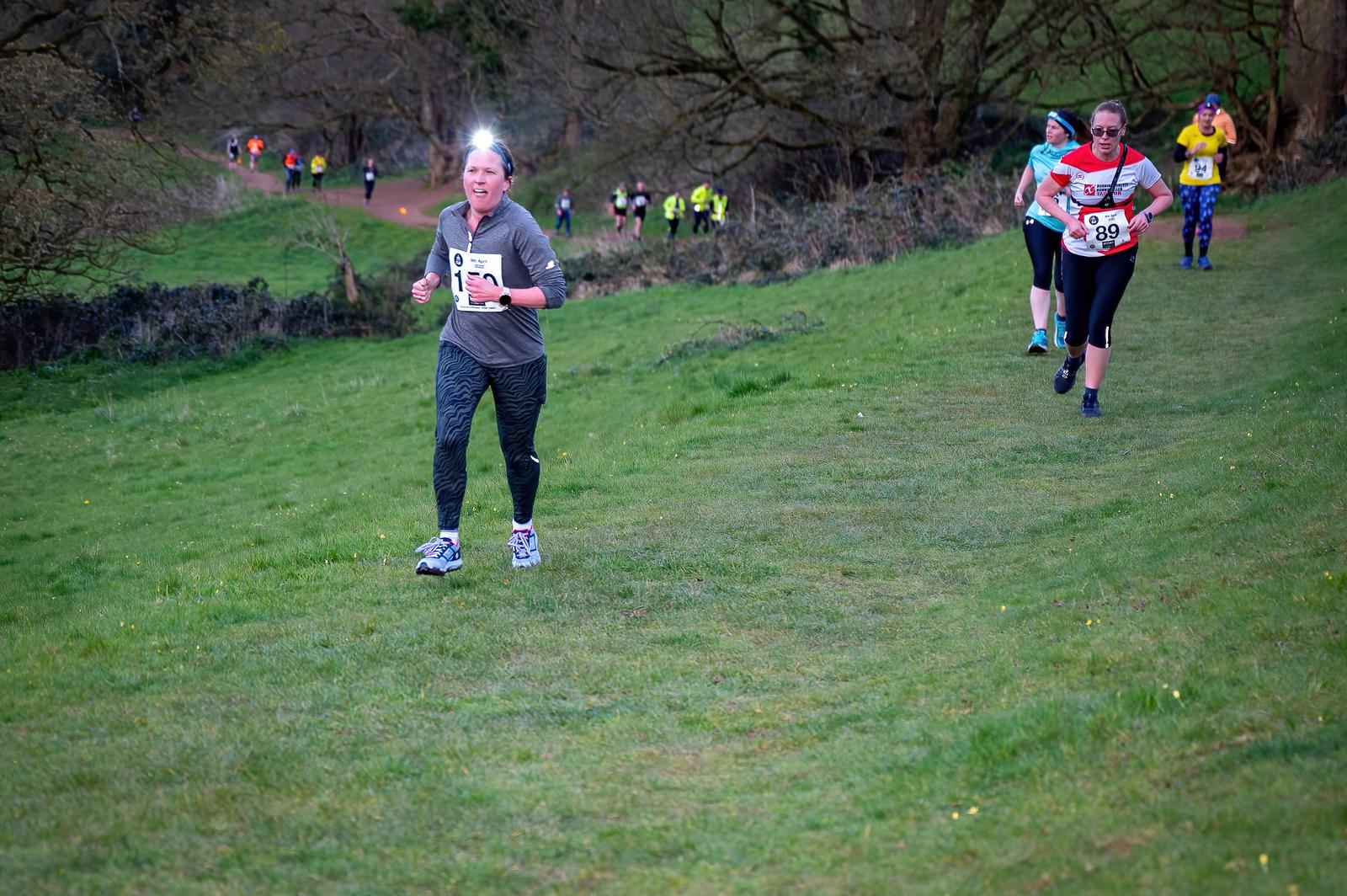 A group of runners participating in a race on a grassy trail. The leading runner is an older woman wearing a grey shirt and black leggings with a headlamp, followed by other runners in colorful athletic wear. Trees and more participants are visible in the background.