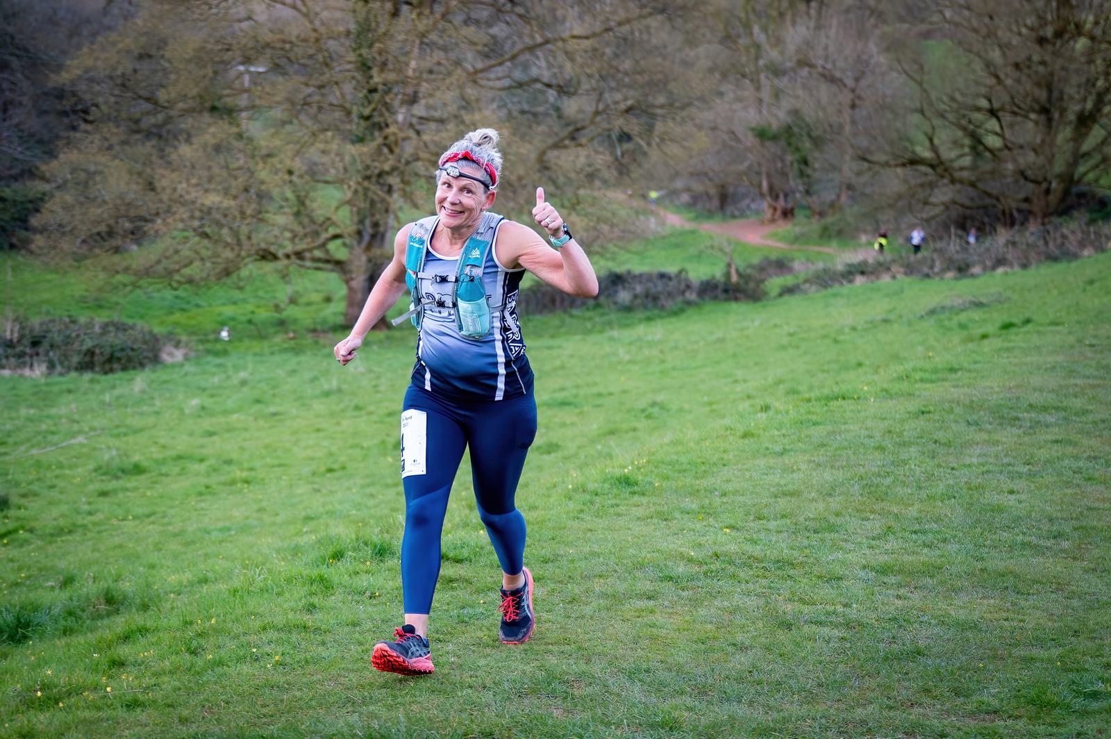 A person in athletic gear runs on a grassy trail, smiling and giving a thumbs-up. They are wearing a red headband, a gray top, and blue leggings with a race number attached. The background features trees and an overcast sky.