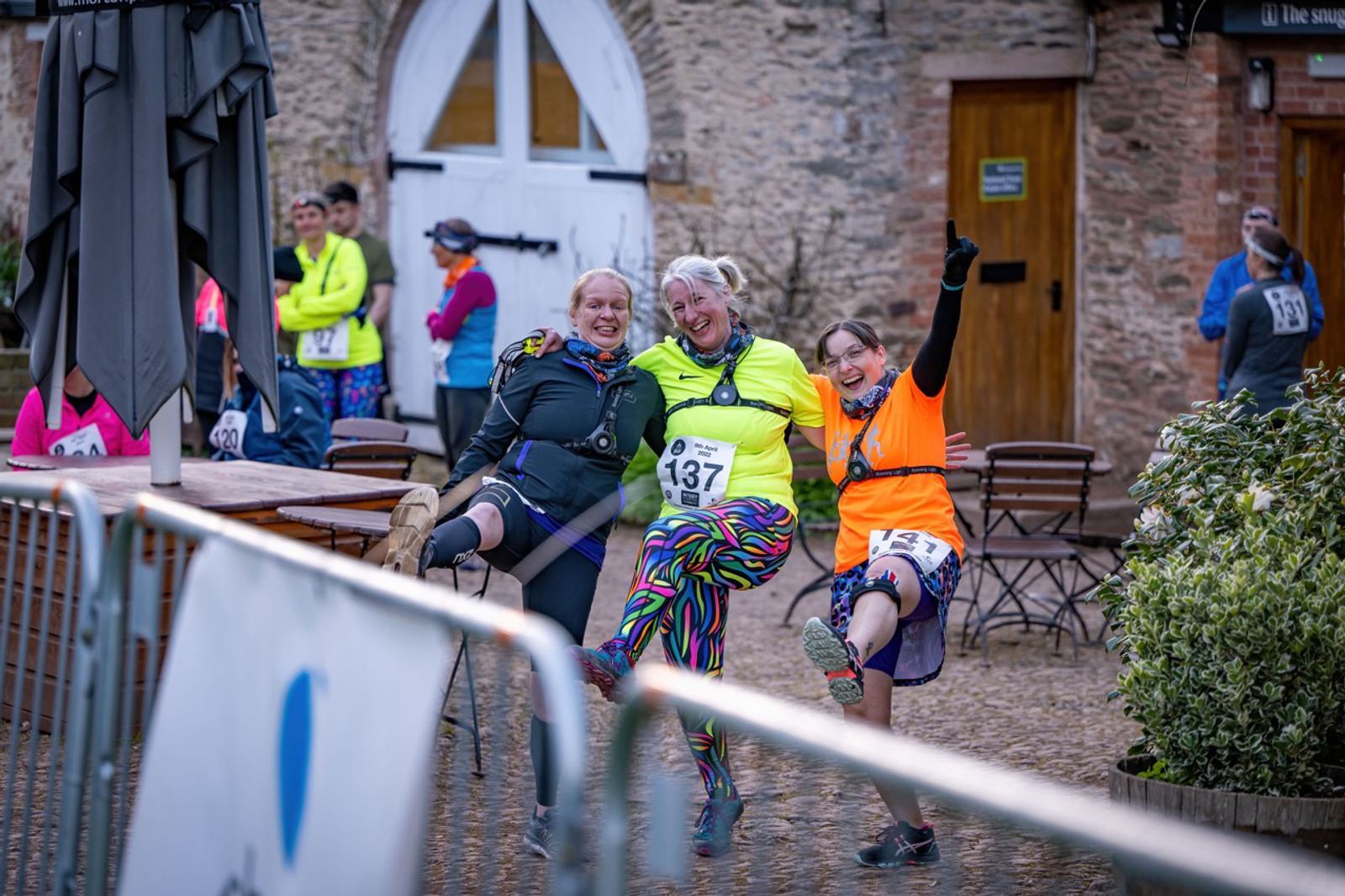 Three women in running gear pose gleefully while lifting one leg, showing their race bib numbers 137, 126, and 141. They're outdoors, next to a metal fence and in front of a stone building with a wooden door. Other runners and spectators are in the background.