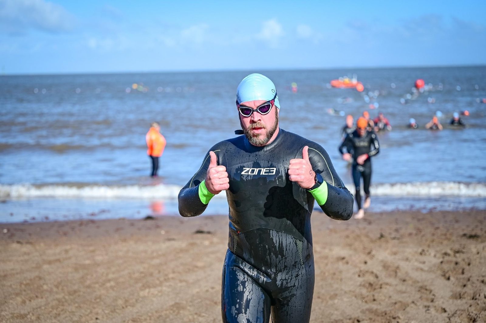 A person wearing a swim cap, sunglasses, and a wetsuit is standing on a beach with their thumbs up. Other swimmers in wetsuits are in the water and along the shoreline in the background. The sky is clear and the ocean is calm.