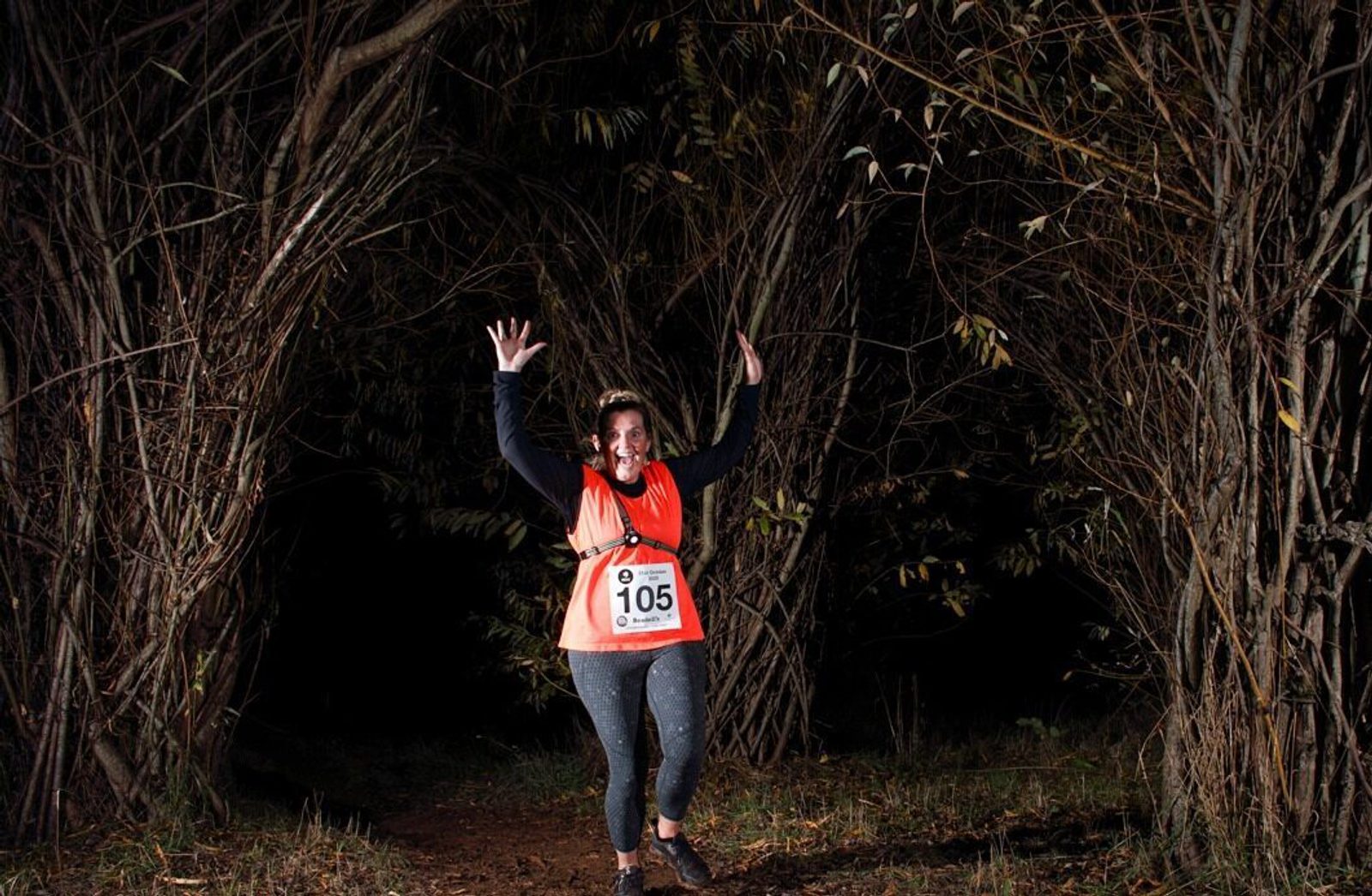 A woman wearing a race bib with the number 105 is running through a forest path at night, illuminated by a spotlight. She is smiling and raising her hands in excitement. The path is surrounded by dense, leafless trees, and event logos are visible at the bottom.