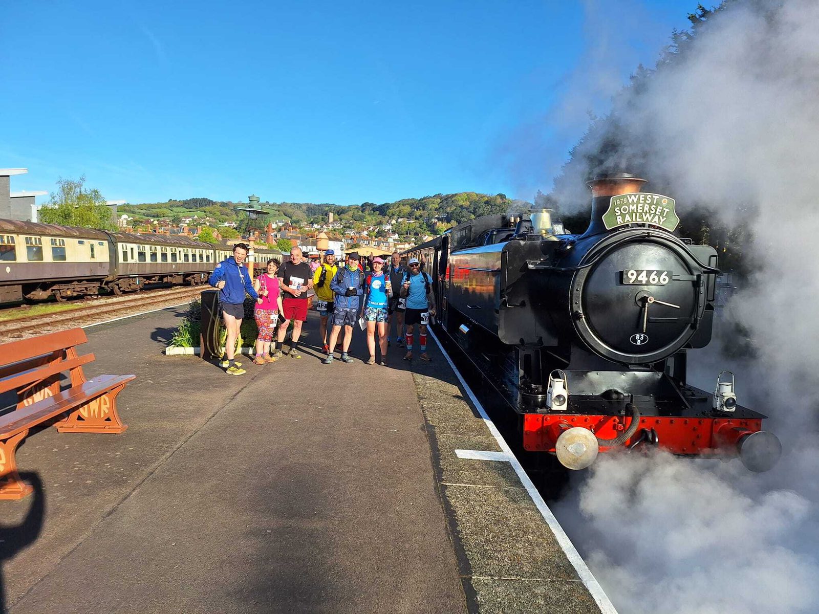A group of people stand on a train platform next to a black steam locomotive with the number 9466 on the front. The platform has a brown bench and is surrounded by scenic hills and greenery under a clear blue sky. Smoke billows from the train's chimney.
