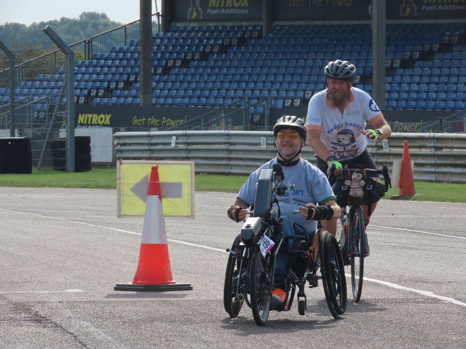 Two cyclists on a track, one using a handcycle and the other on a standard bike. The background features empty blue stadium seats and safety cones. Both cyclists wear helmets, and the track is marked with signage and barriers.